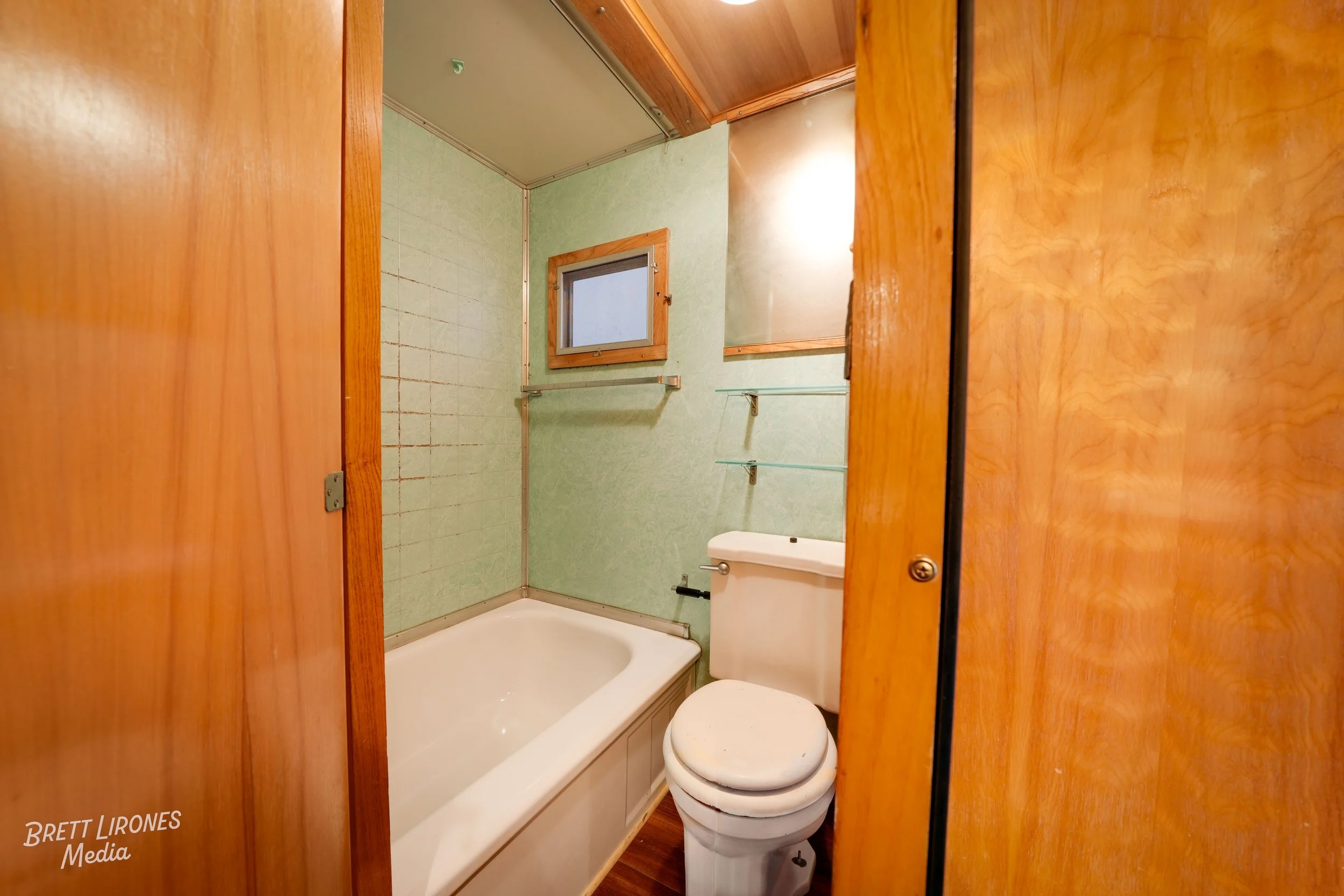 Small bathroom with a bathtub, toilet, and wooden walls and door, with a small window and shelves on the wall.