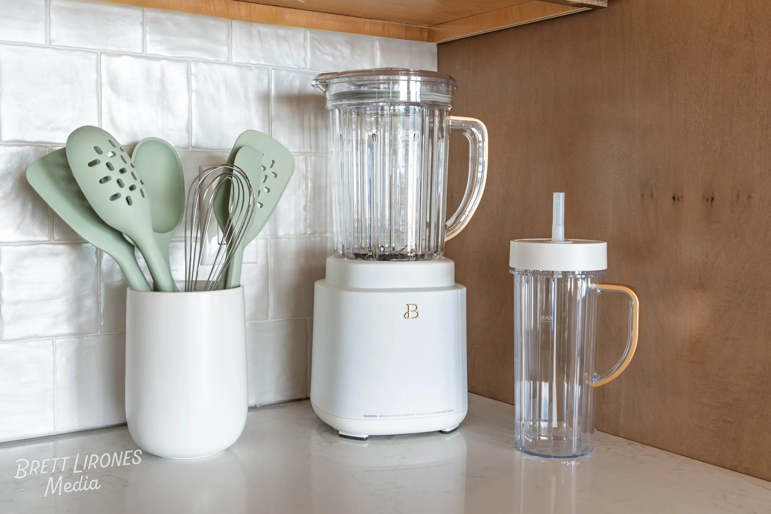 Kitchen counter with a white container holding green silicone spatulas and a metal whisk, a white blender with a clear pitcher, and a clear tumbler with a white lid and straw, all against a tiled white backsplash and wooden wall.