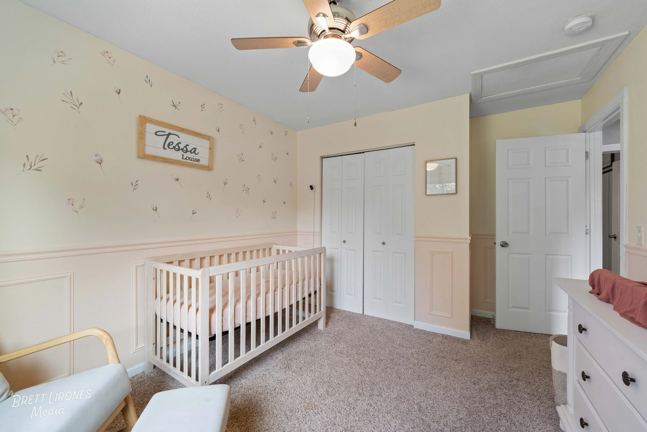 Nursery room with a white crib, a white dresser with a changing pad, a ceiling fan with wooden blades, beige carpet, and cream walls decorated with small floral designs. A sign above the crib reads 'Tessa Louise'.