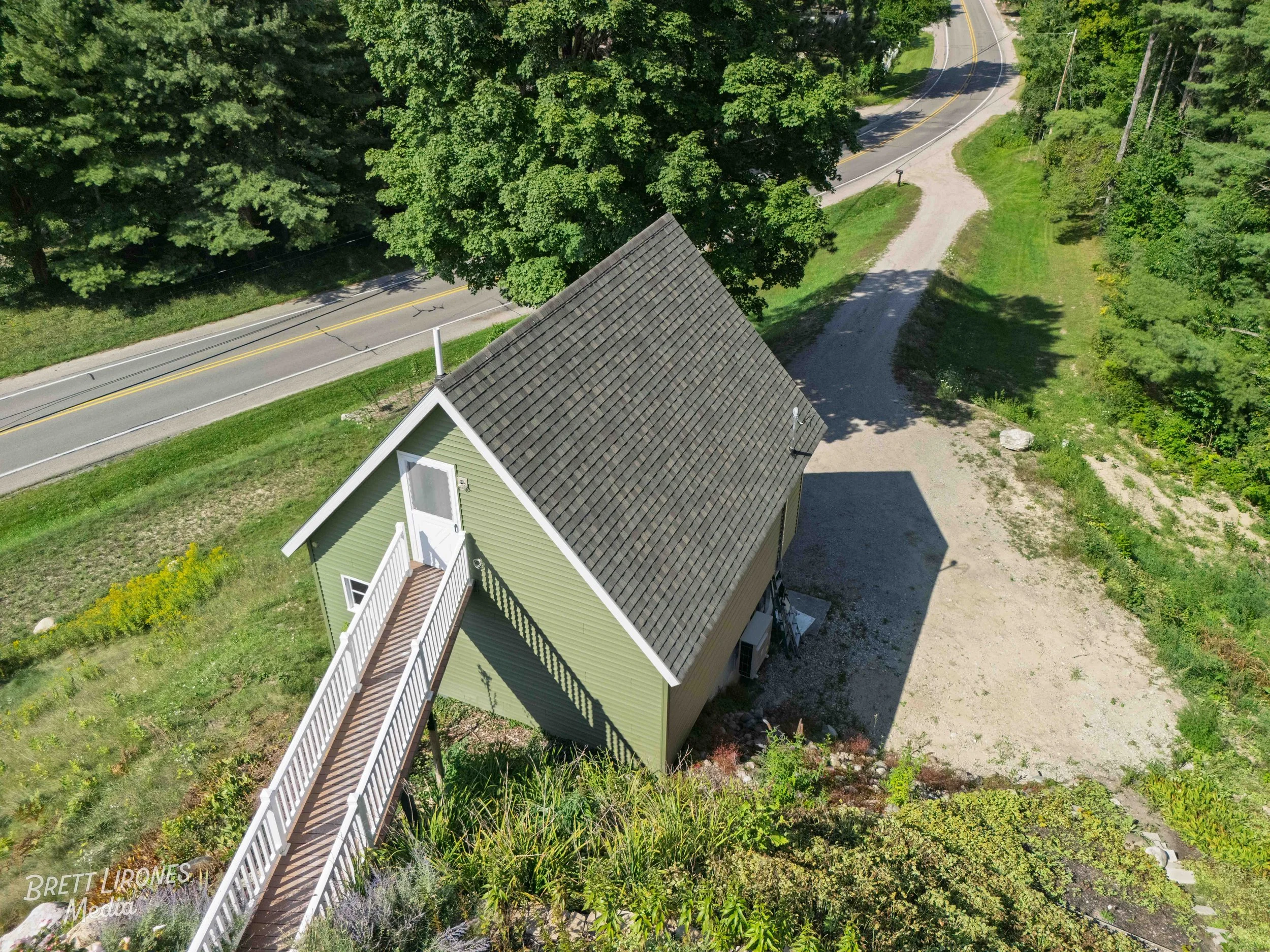 An aerial view of a green house with a pitched roof, connected to a raised walkway leading up to a door. The house is surrounded by trees and grass, with a gravel driveway and a paved road nearby.