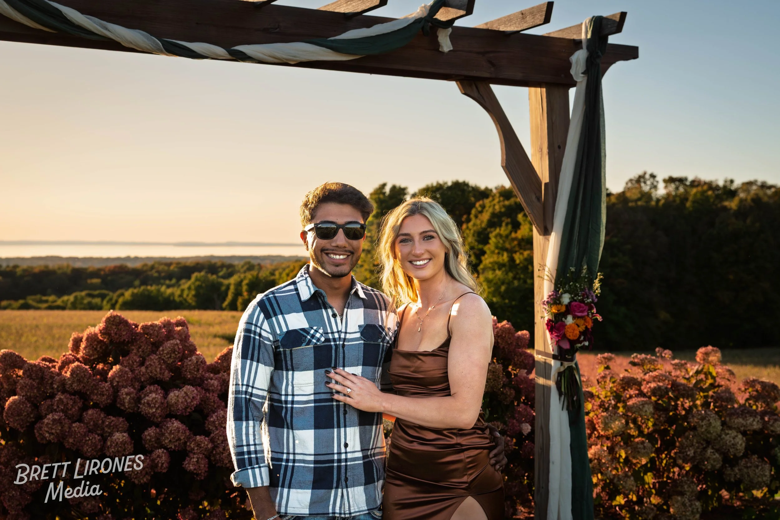 A smiling couple standing under a wooden arch decorated with fabric and flowers, during a sunset outdoors. The man is wearing sunglasses and a plaid shirt, and the woman is in a brown dress with her arm around the man's waist, with a wedding or roman