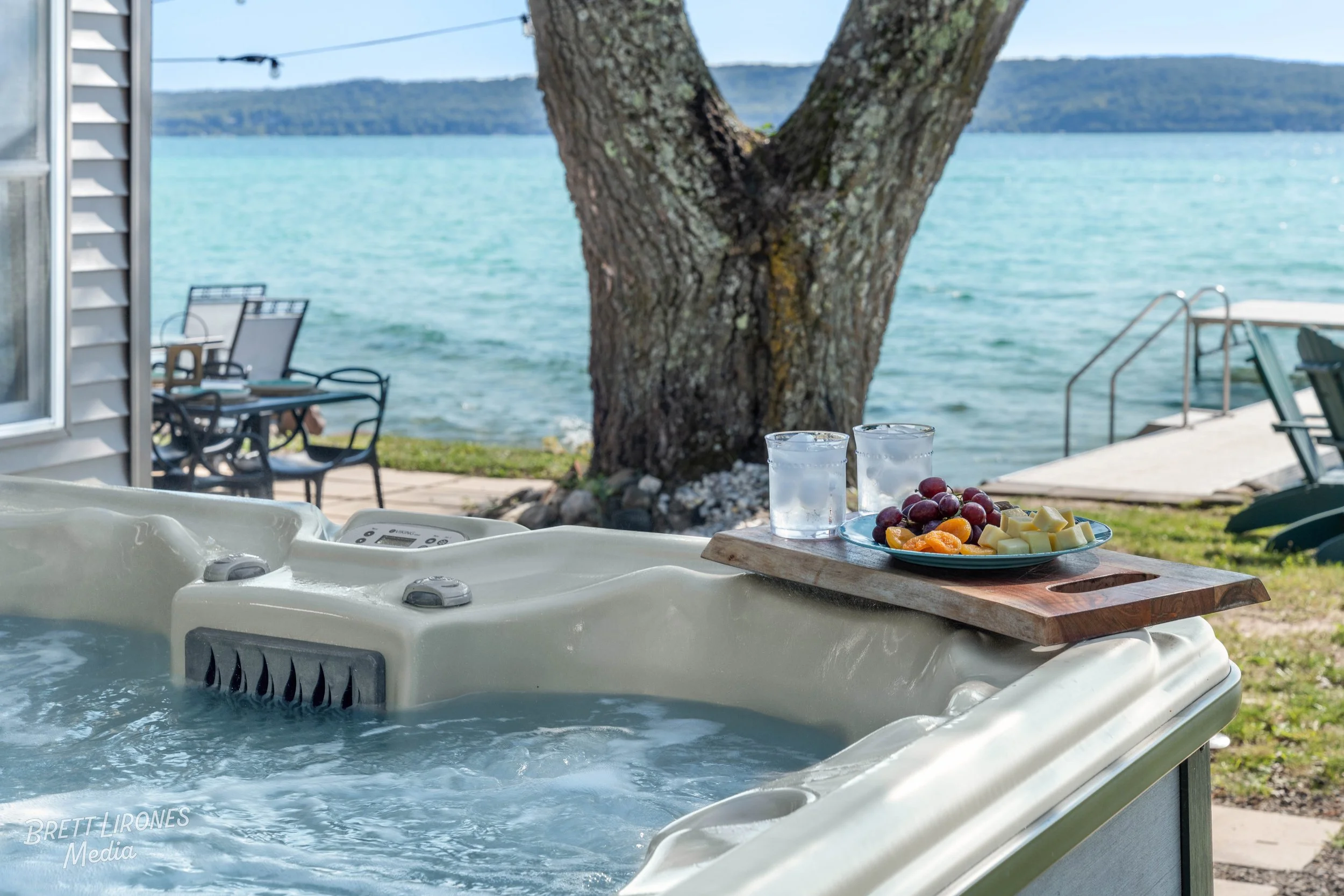 Hot tub with a wooden tray holding two glasses of water and a plate of grapes, cheese, and fruit, set outdoors by a lake with a large tree, patio furniture, and a dock in the background.