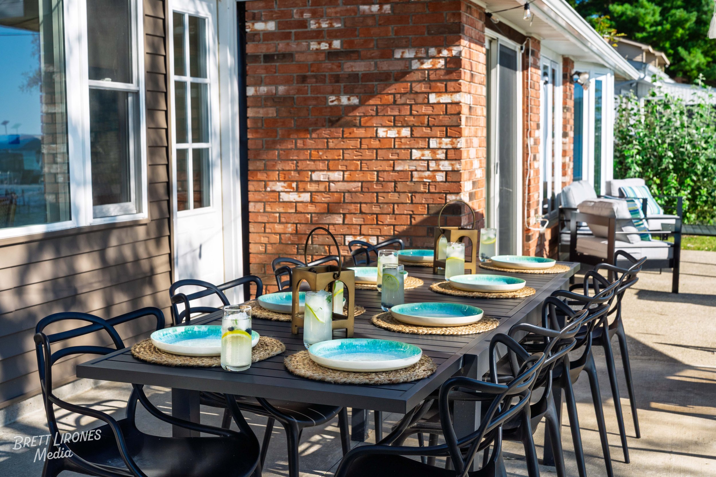 Outdoor dining table set with seven turquoise ceramic plates on woven placemats and glasses of lemonade, placed on a patio next to a brick house with sliding glass doors and patio furniture in the background, under a sunny sky.