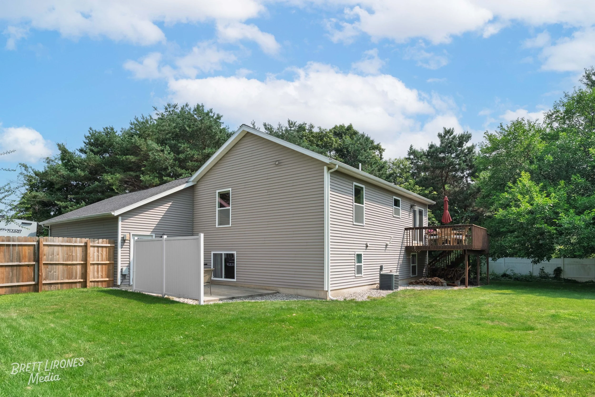 Rear view of a two-story house with beige vinyl siding, a wooden deck with a red umbrella, a grassy backyard, surrounded by trees and a wooden fence.