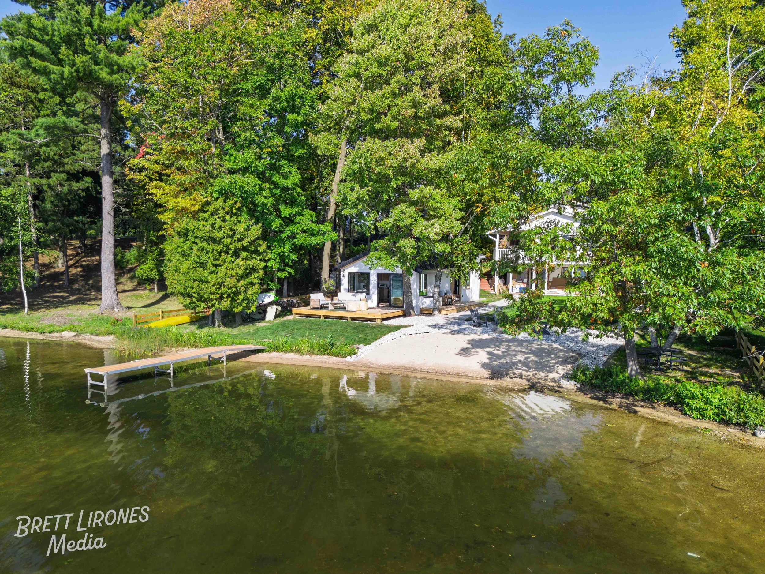 A lakeside property with a house surrounded by trees, featuring a dock extending into the water and a small sandy beach area.