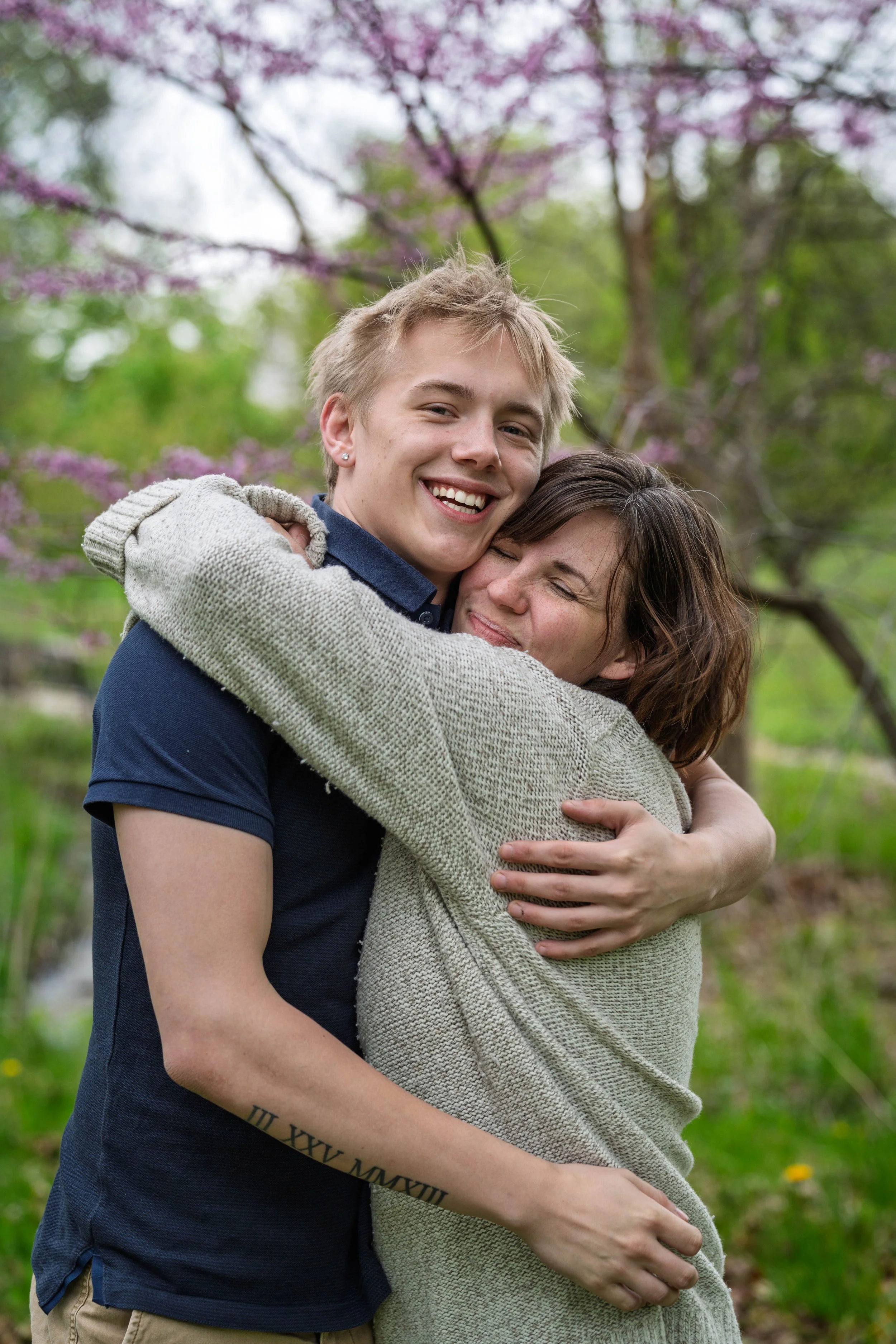 A young man and woman are happily hugging outdoors with pink flowering trees in the background.