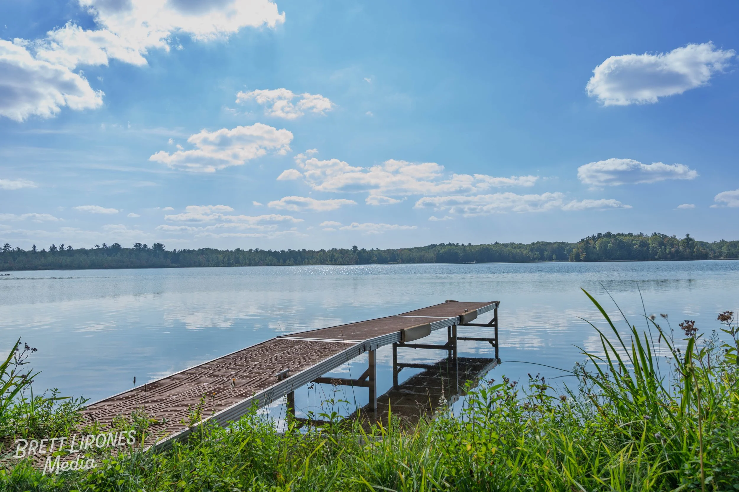 A wooden dock extends from the grassy shoreline into a calm lake under a partly cloudy sky with scattered clouds.