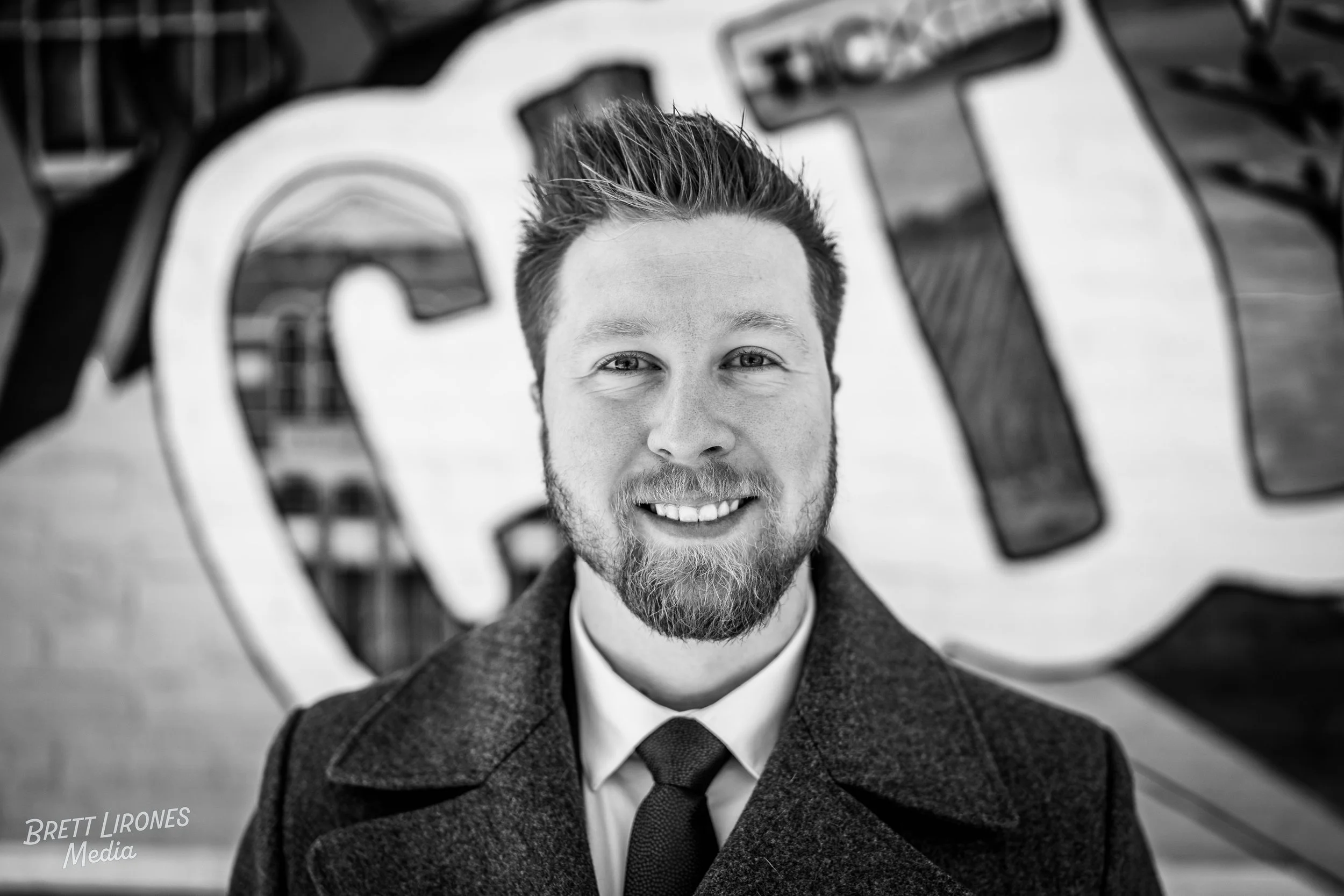 Black and white photo of a young man with a beard, smiling, wearing a coat and tie, standing in front of a graffiti wall.