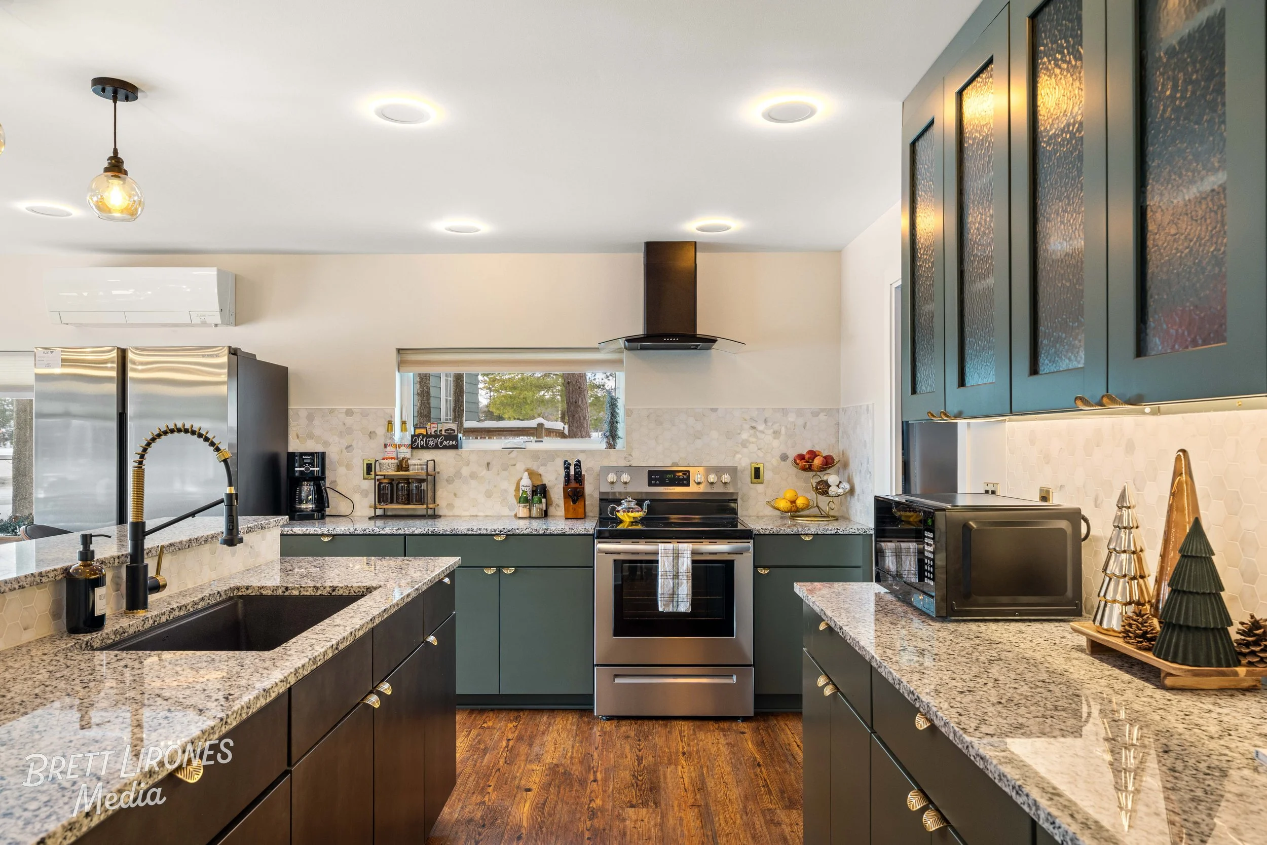 Modern kitchen with gray and teal cabinets, stainless steel appliances, granite countertops, and wood flooring, decorated for the holidays with small Christmas trees and pine cones.