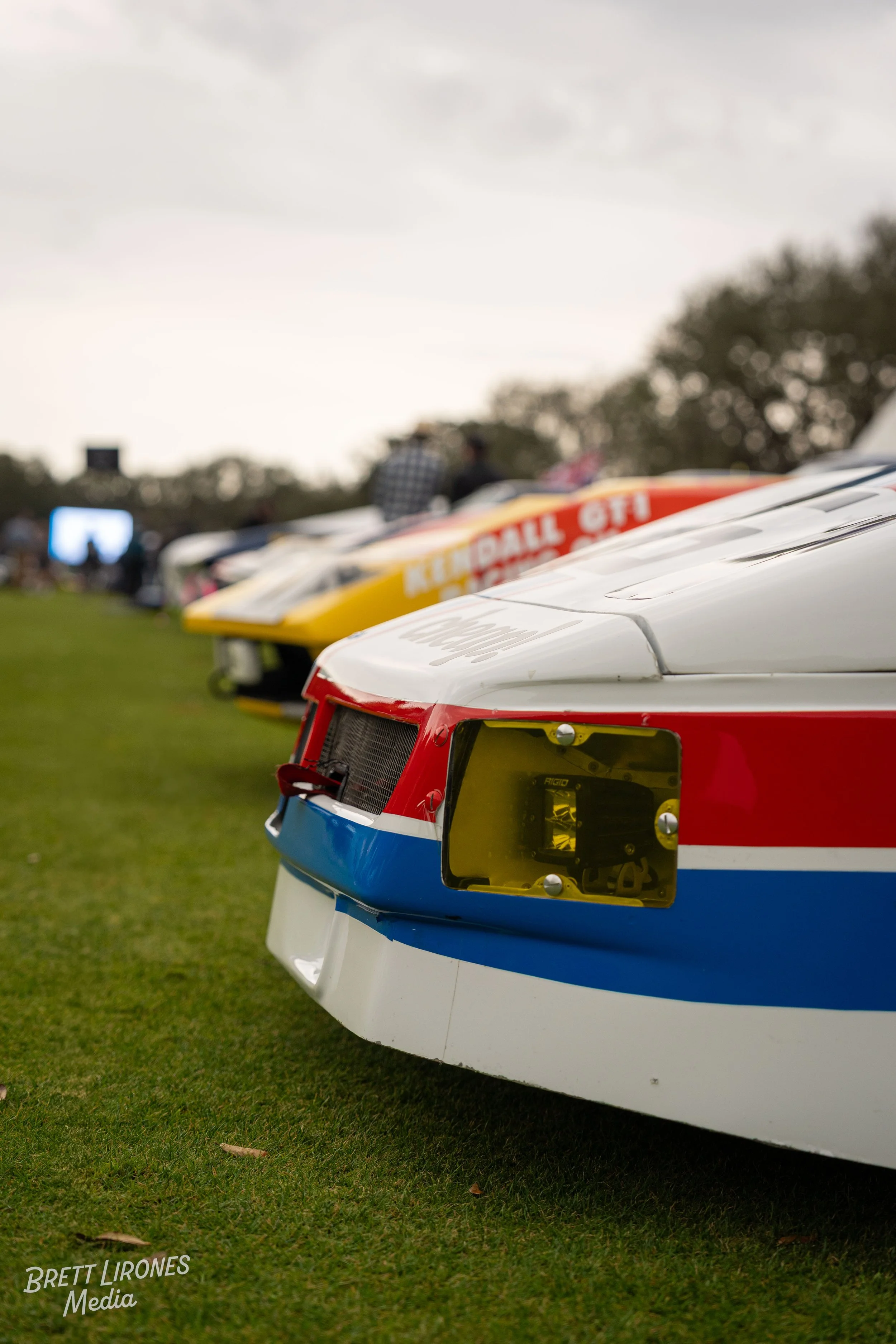 Close-up of race cars lined up on grass field, with the front of a car featuring blue, red, and white colors in focus, and others in the background slightly blurred.