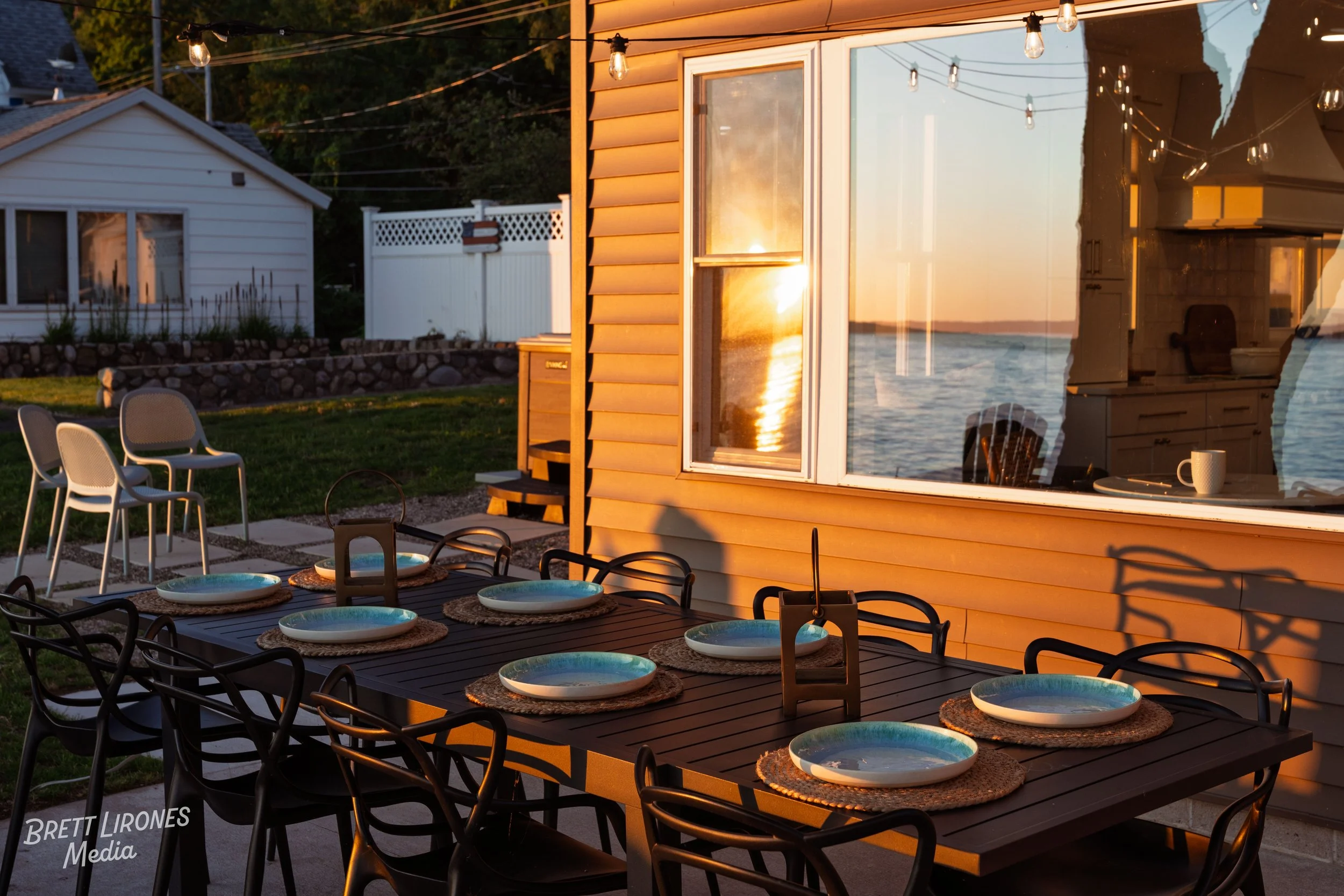 Outdoor dining table set with plates and placemats in evening sunlight on a patio by a lake, with string lights overhead and a reflection of the sunset in the window of a yellow house.