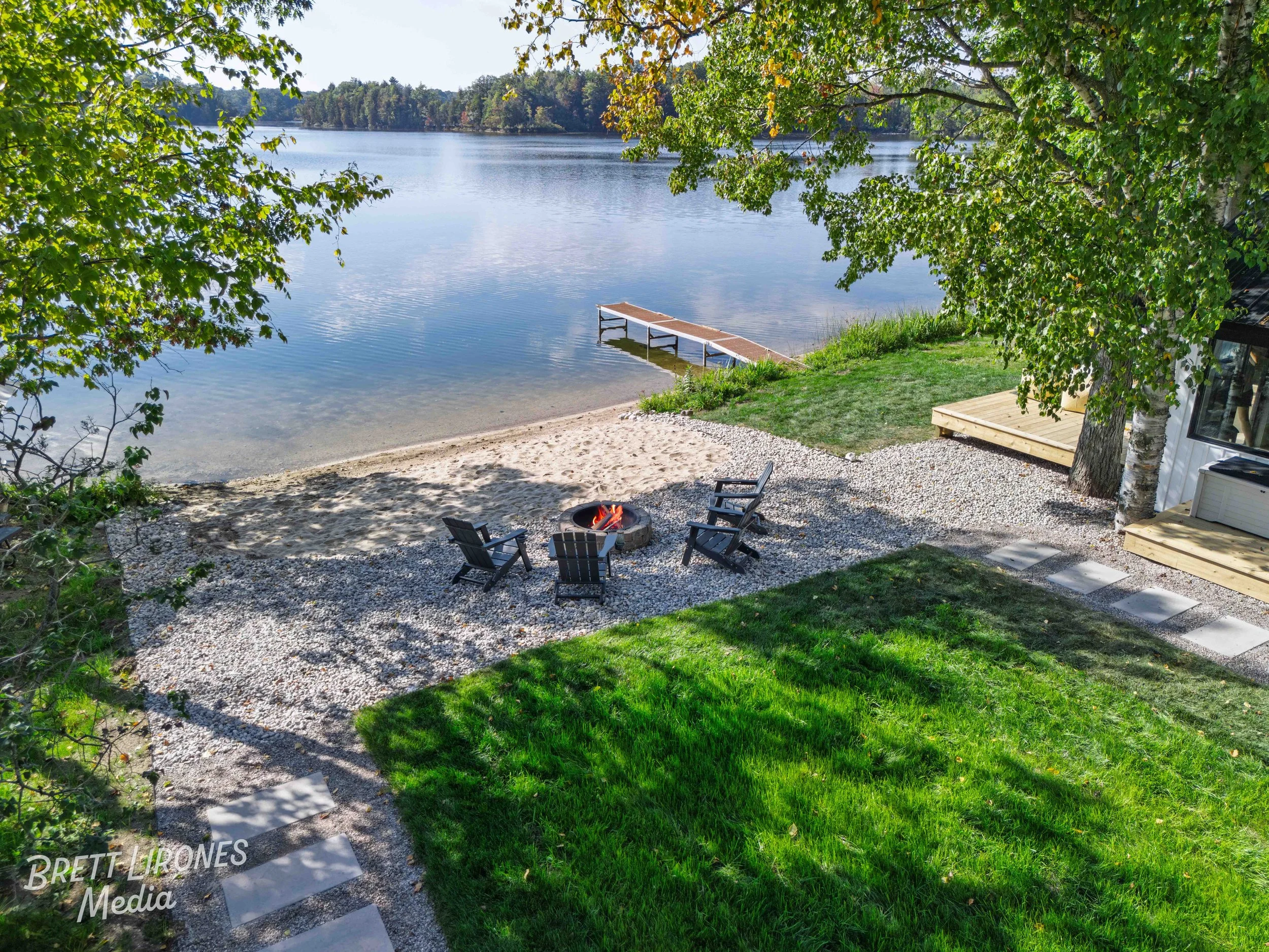 A lakeside outdoor area with four chairs arranged around a fire pit, a sandy beach, a small pier extending into the water, and surrounded by trees and grass.