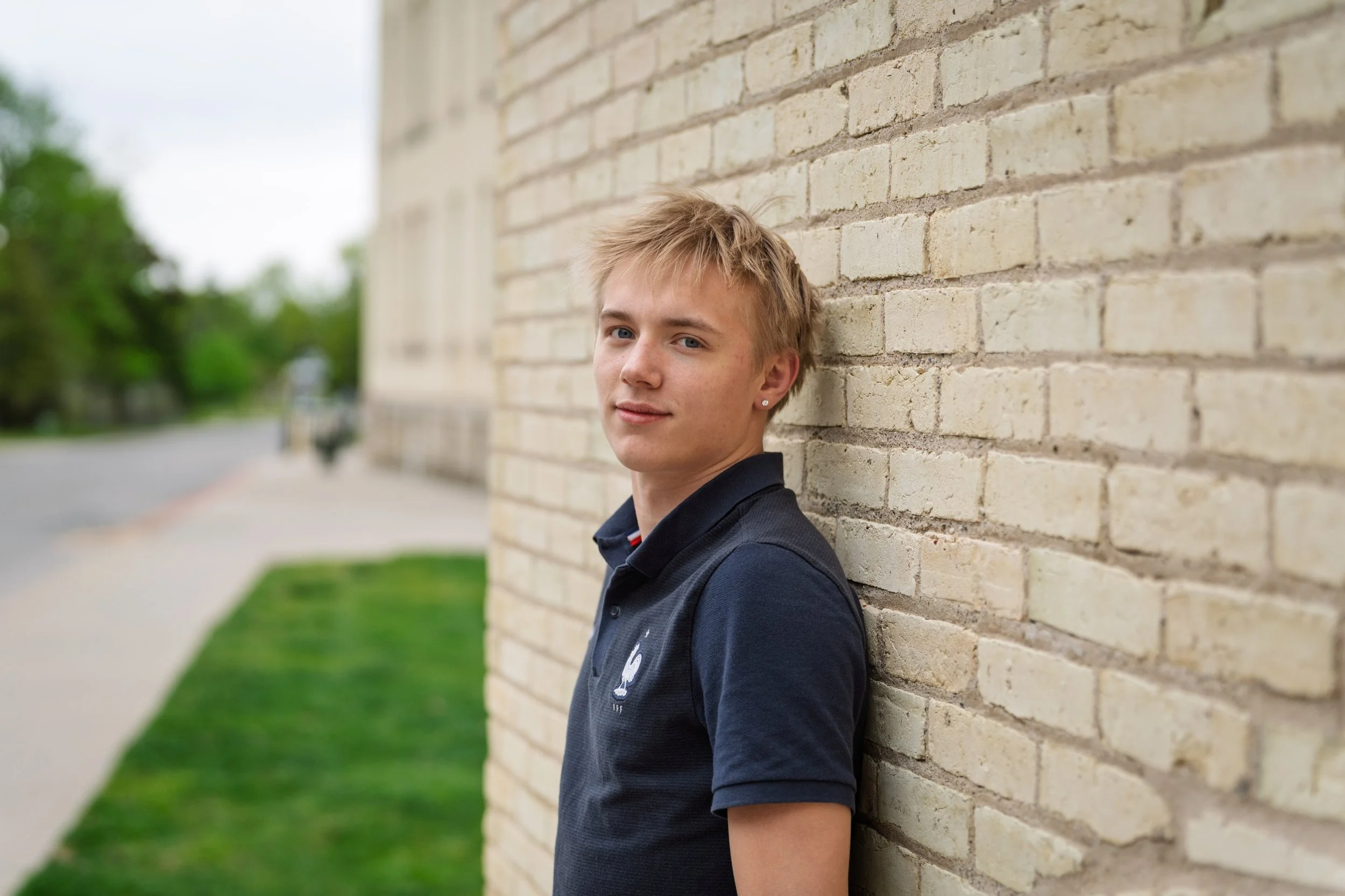 Young man leaning against a brick wall outdoors, with a blurred background of a sidewalk, grass, and trees.