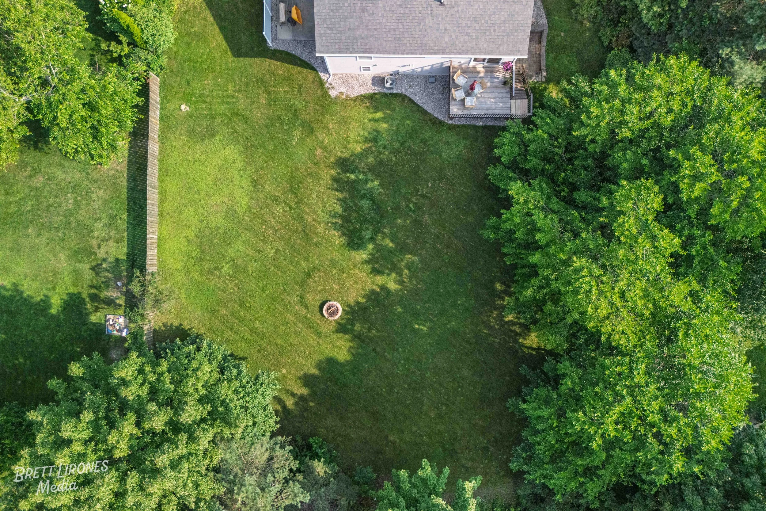An aerial view of a backyard with a grassy lawn, a fire pit in the center, a wooden deck with outdoor furniture attached to a house, and large trees surrounding the yard.
