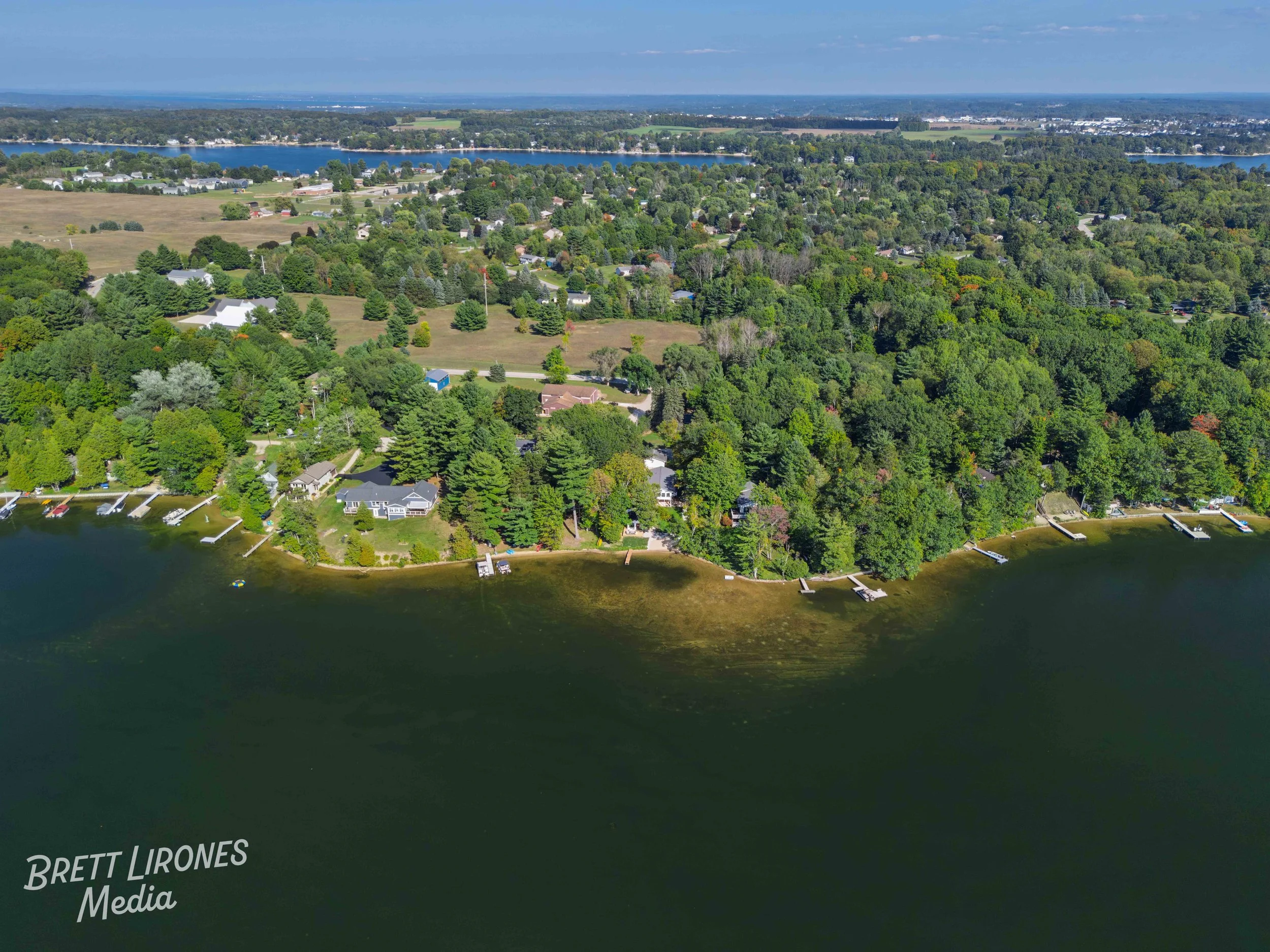 Aerial view of a lakeside neighborhood with scattered houses, docks, and lush green trees along the water's edge, with a mix of forested and open land in the background under a partly cloudy sky.