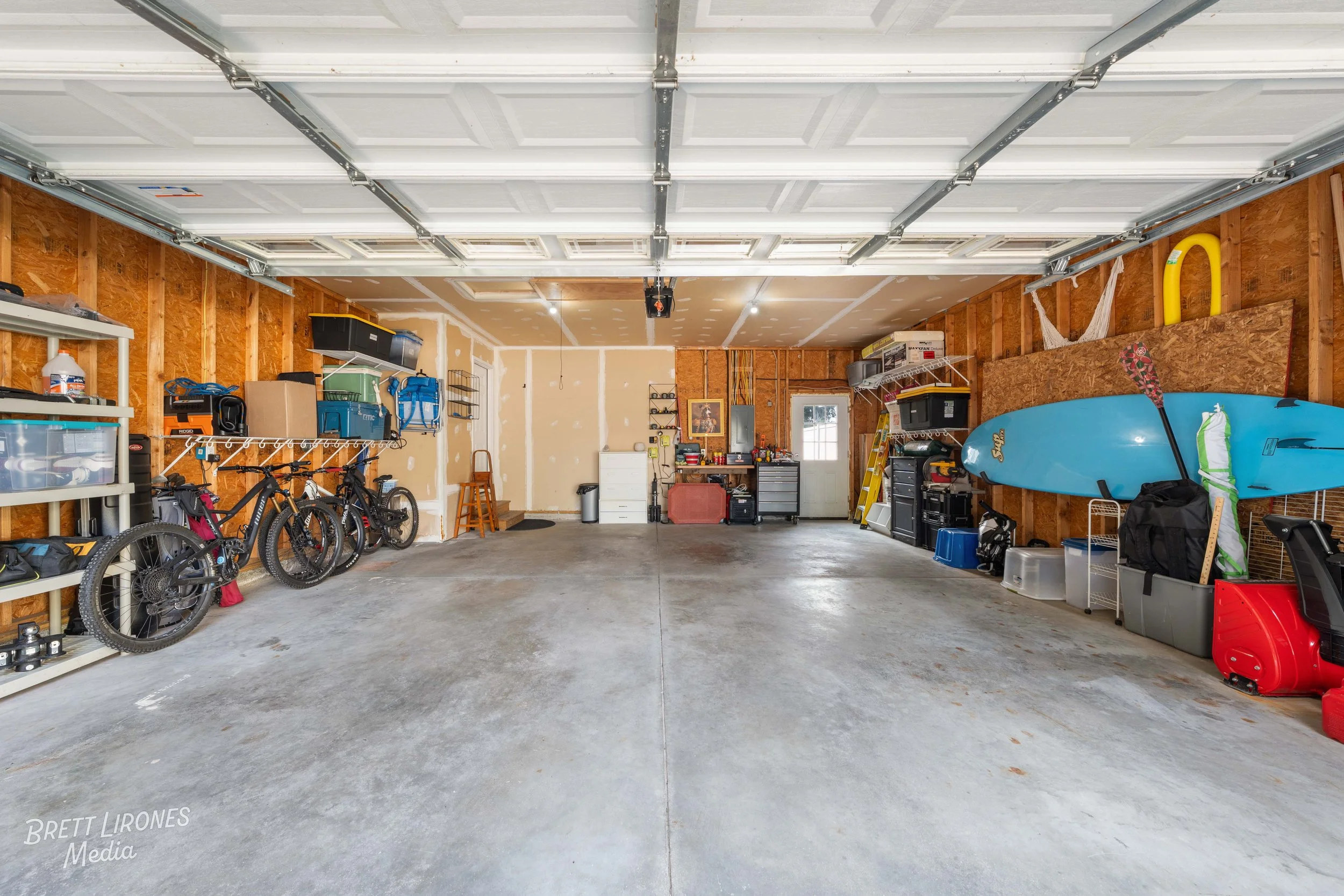 Empty garage with bicycles leaning against the left wall, various tools and storage containers along the walls, a surfboard hanging on the right wall, and a table with more tools and equipment at the back. The garage has a partly finished interior wi
