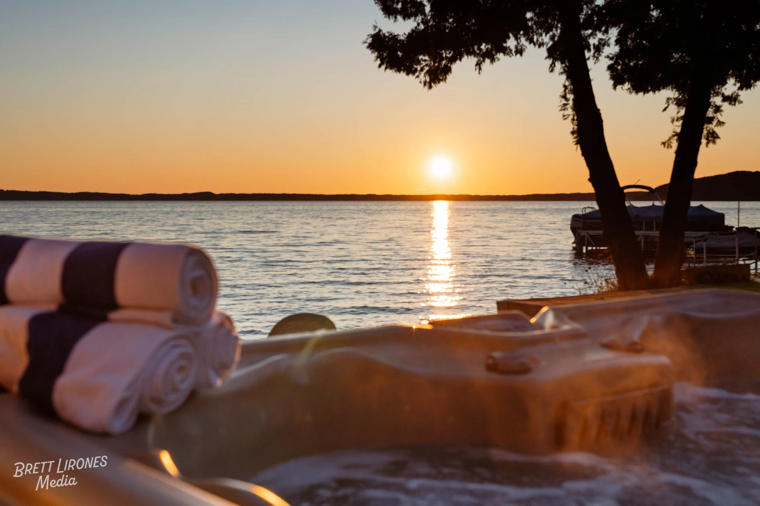 Hot tub with rolled towels on the edge, overlooking a sunset over the water with boats and trees in the background.