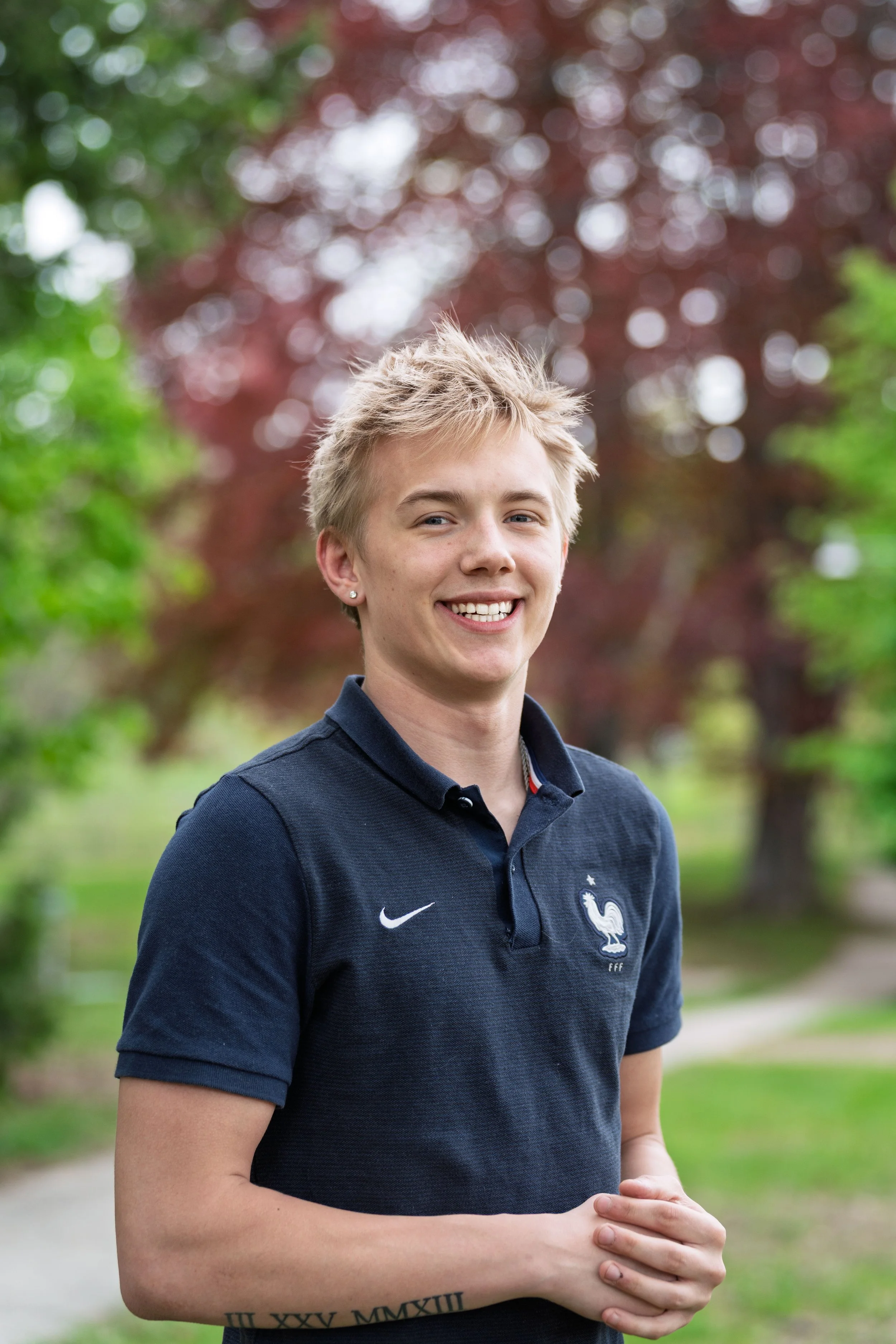 Young woman with short blonde hair smiling outdoors, wearing a navy blue polo shirt with the French soccer team logo and Nike swoosh, standing in a park with green and red trees in the background.