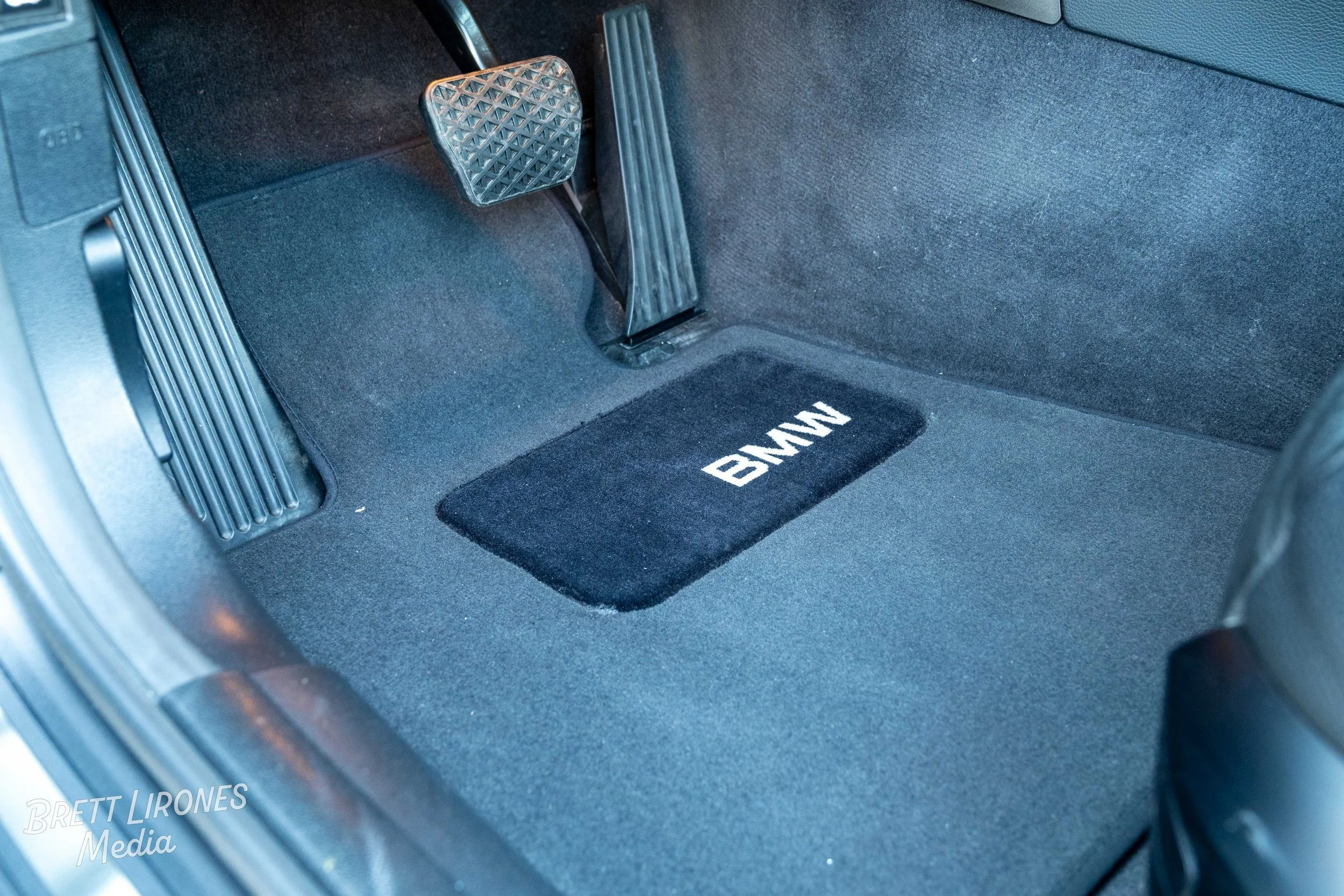 The image shows the interior floor of a BMW vehicle, including the accelerator pedal, brake pedal, and a black floor mat with the BMW logo embroidered in white.