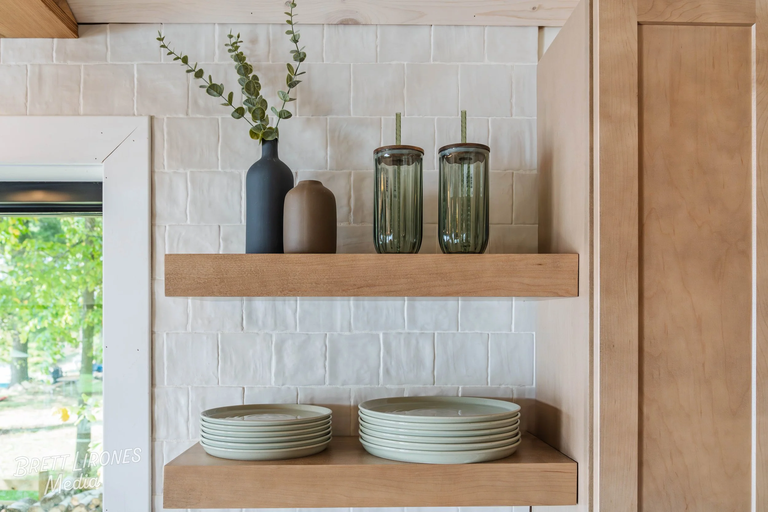 Open kitchen cabinet with two wooden shelves holding green ceramic plates on the lower shelf, two green glass tumblers on the upper shelf, and decorative vases with greenery, against a white brick wall with a window showing greenery outside.