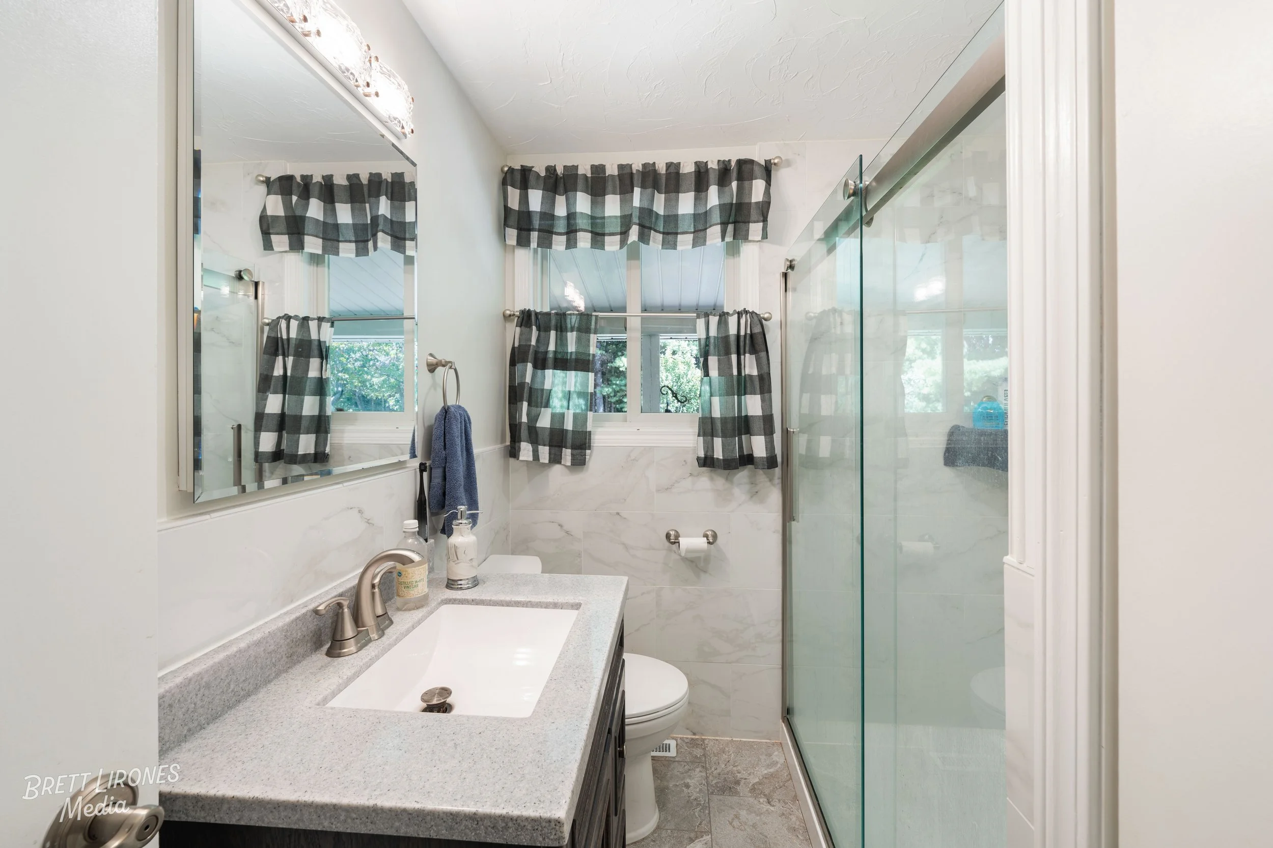 Bathroom with white walls, marble tiles, a glass shower door, a vanity with a sink and a large mirror. There are checkered curtains on the window, a towel ring with a blue towel, and various toiletries on the vanity counter.