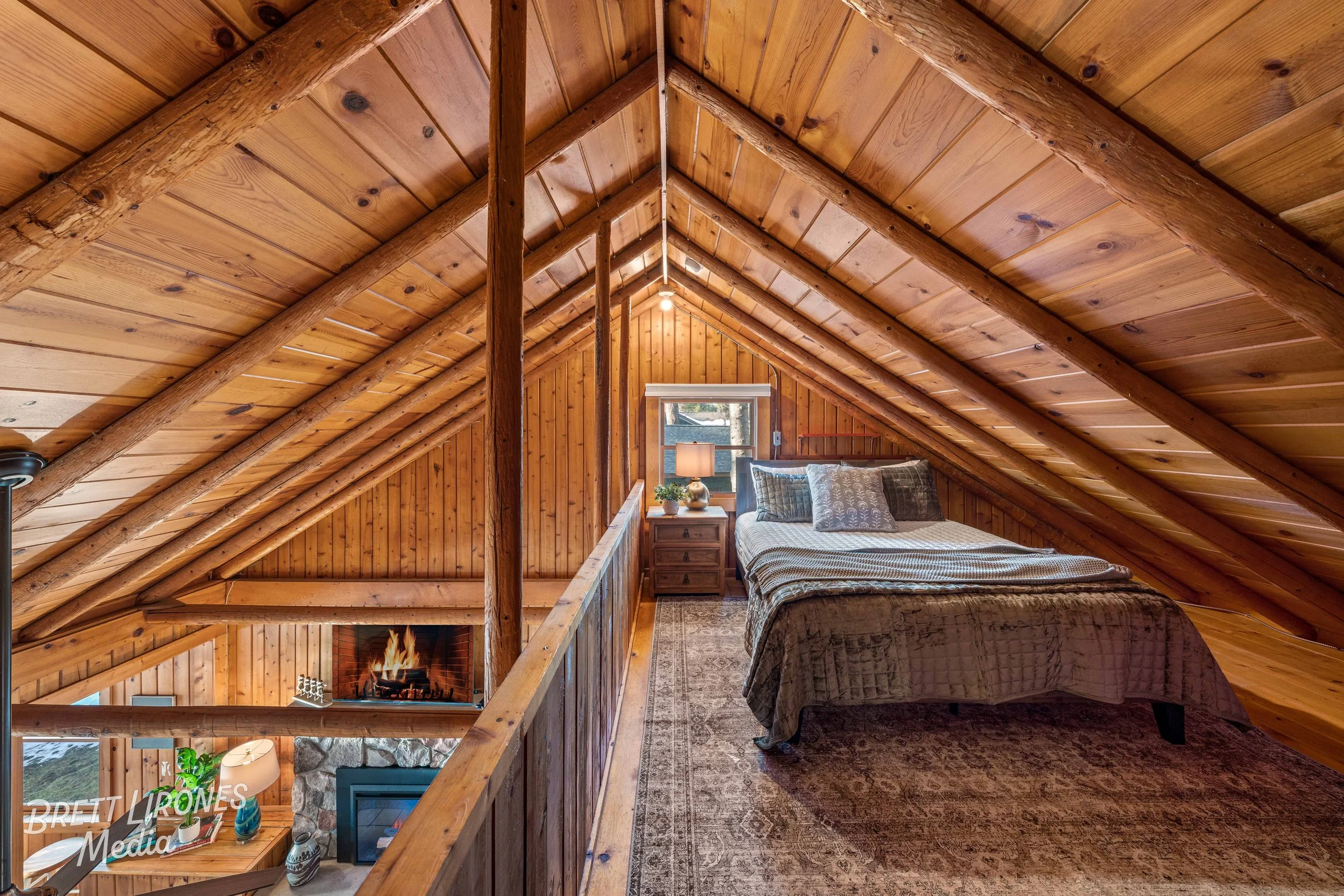 A cozy attic bedroom with wood-paneled walls and sloped ceiling, containing a bed with gray and white bedding, bedside table with lamp, and a small window, overlooking a lower living space with a fireplace.