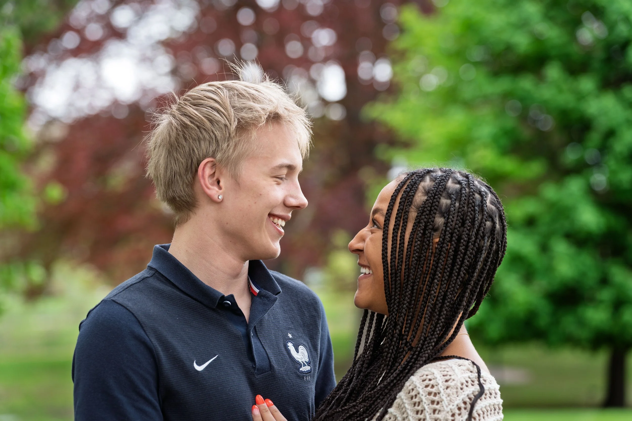 A young man and woman face each other outdoors, smiling and looking into each other's eyes, with a background of green and reddish-brown trees.