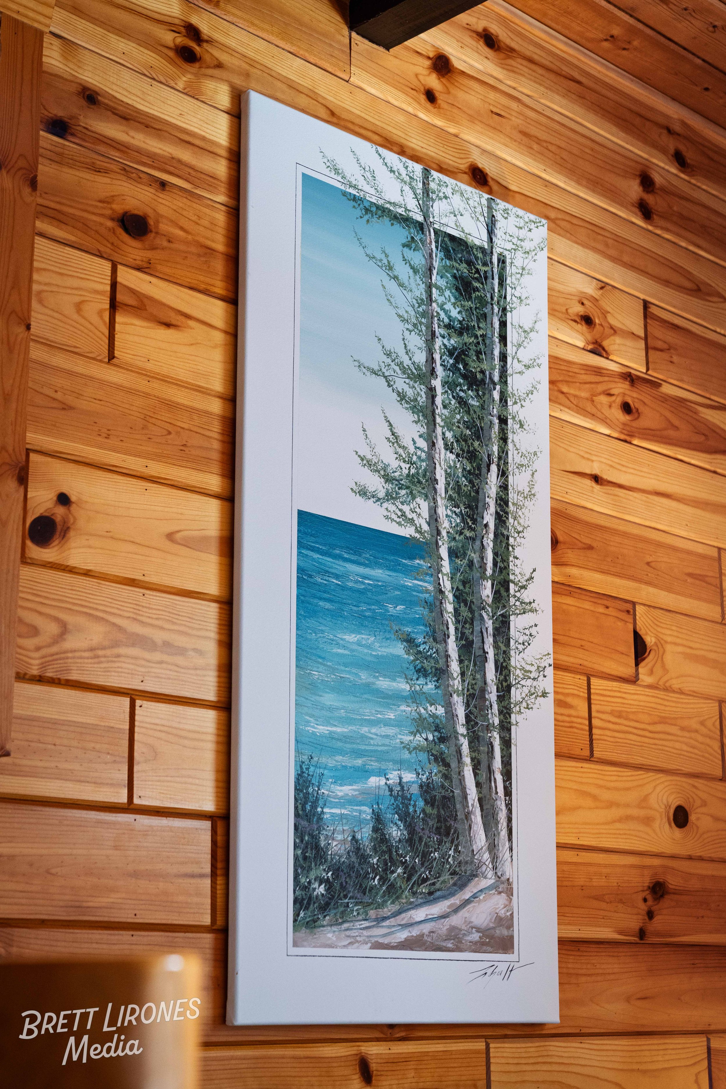 Painting of a coastal scene with trees in the foreground and the ocean in the background, hung on a wooden wall.