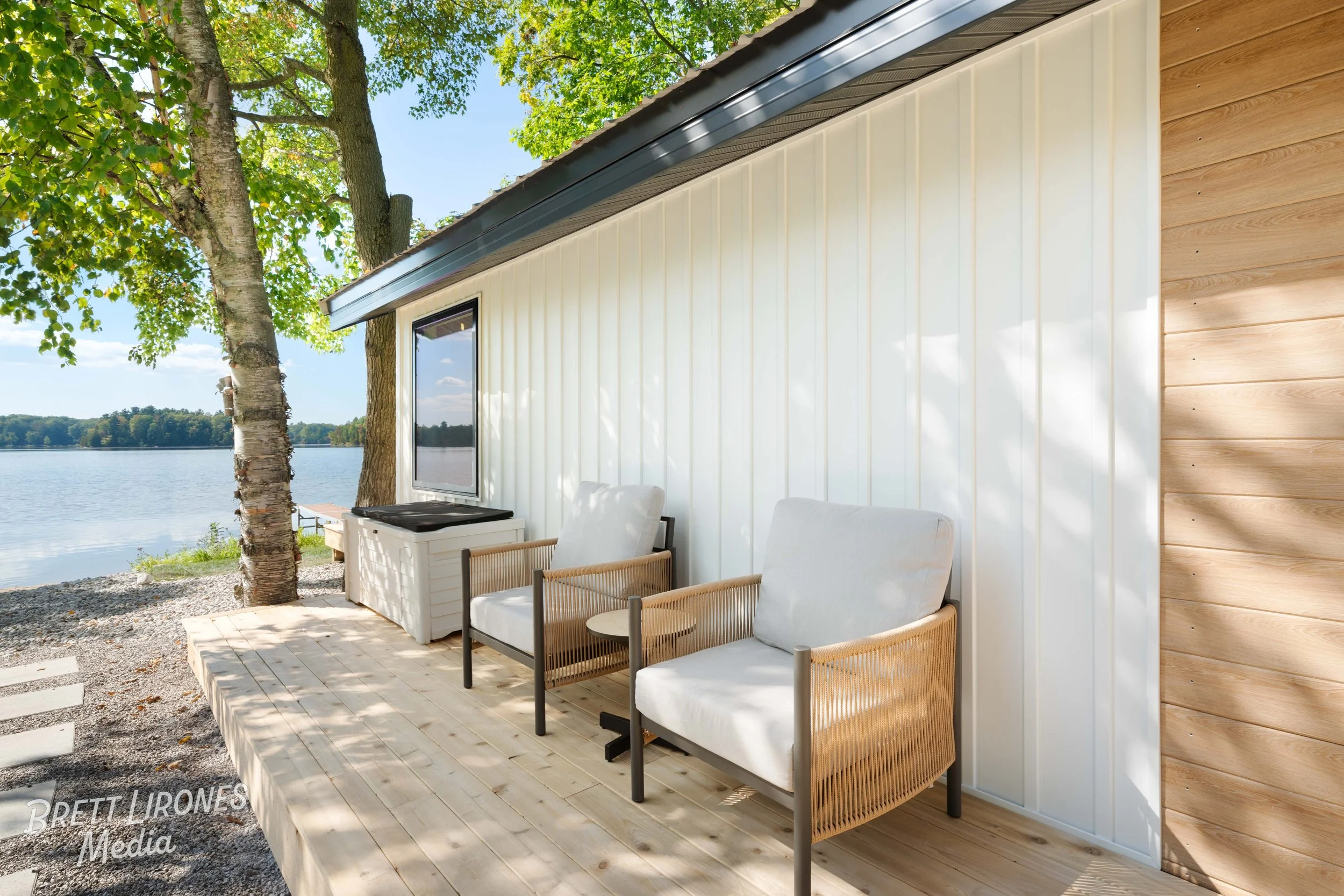 A lakeside patio with two white cushioned chairs and one wooden armchair on a wooden deck, beside a house with white and wood siding, surrounded by green trees and overlooking a calm lake.