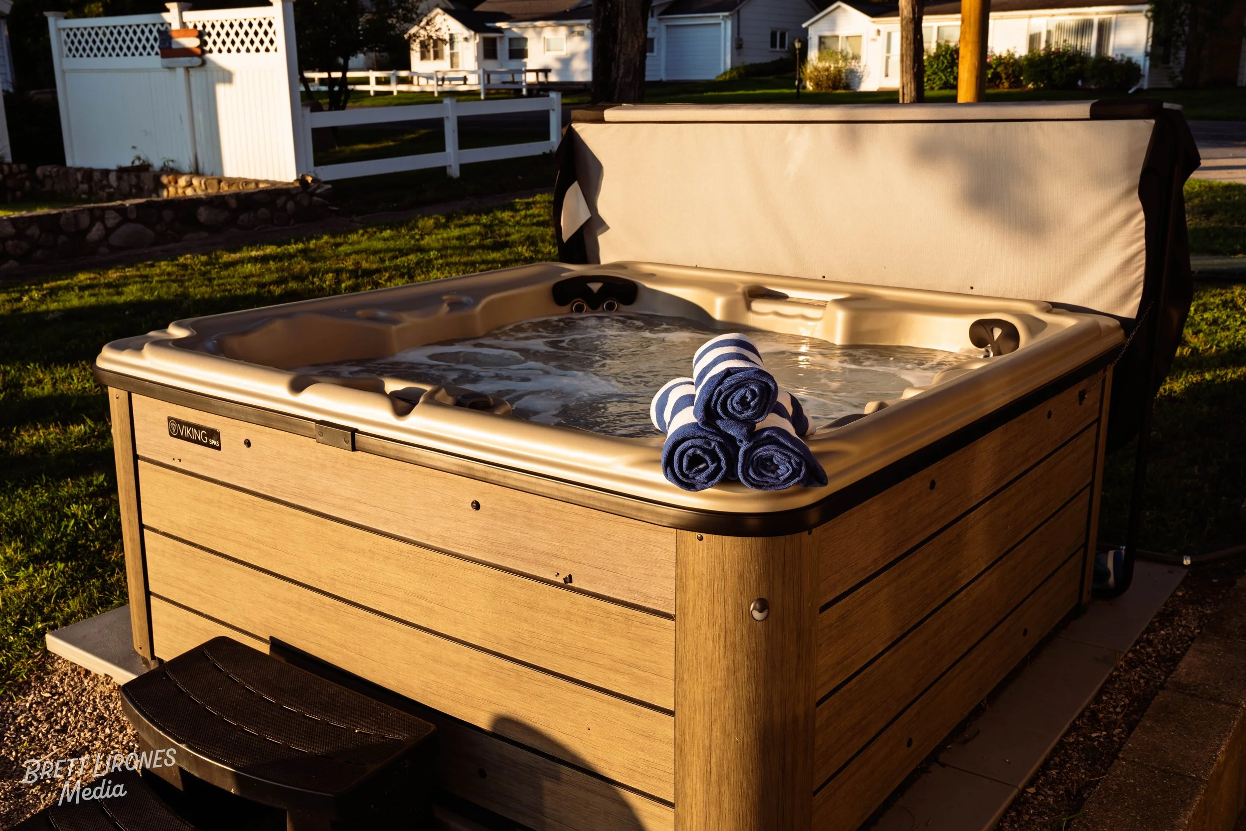 Wooden hot tub filled with water, with three rolled towels on the edge, outdoors in a yard with grass and white fences, in late afternoon sunlight.