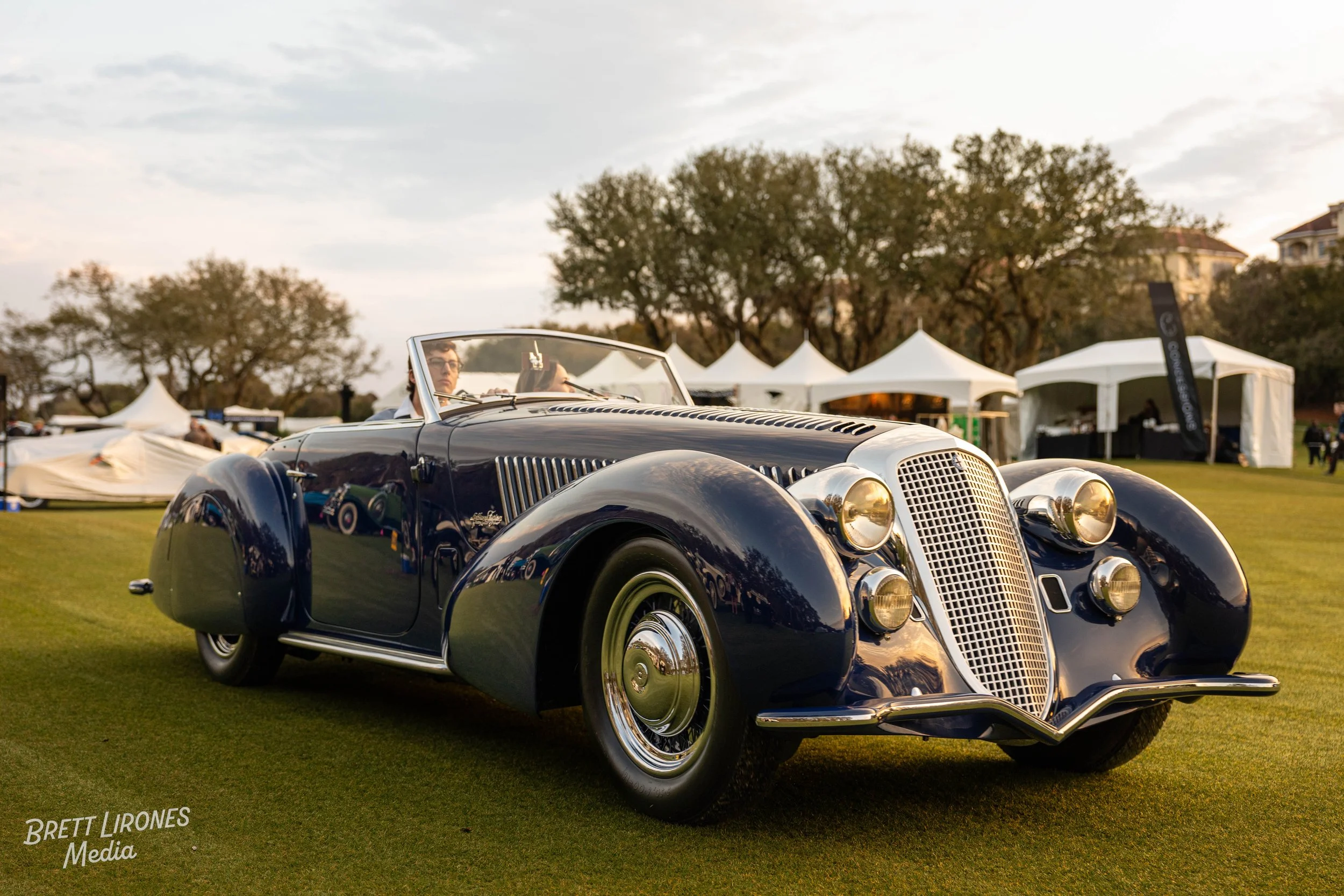 A vintage black luxury car with chrome accents parked on a grassy area at an outdoor event, with white tents and trees in the background during sunset.