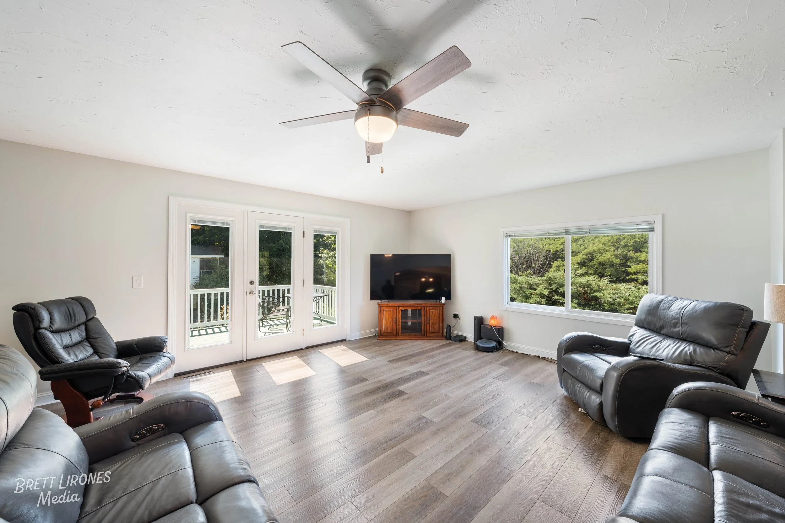 Living room with leather sofas, a TV on a wooden stand, large window, and glass doors leading to a balcony with outdoor seating.