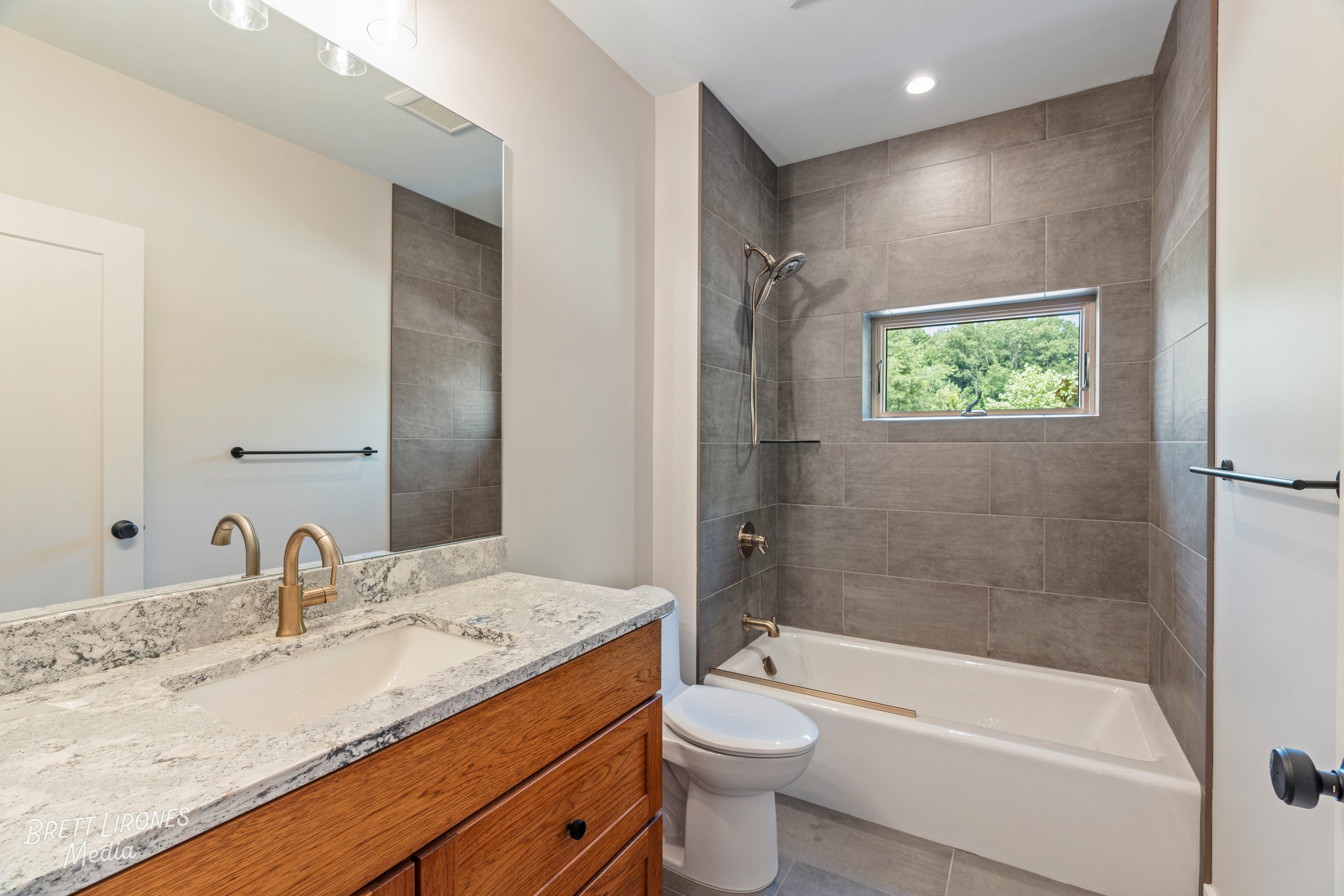 Bathroom with a vanity, sink with a granite countertop, a large mirror, a toilet, a bathtub with a gray tiled wall, a small window showing green trees outside, and black towel bars.