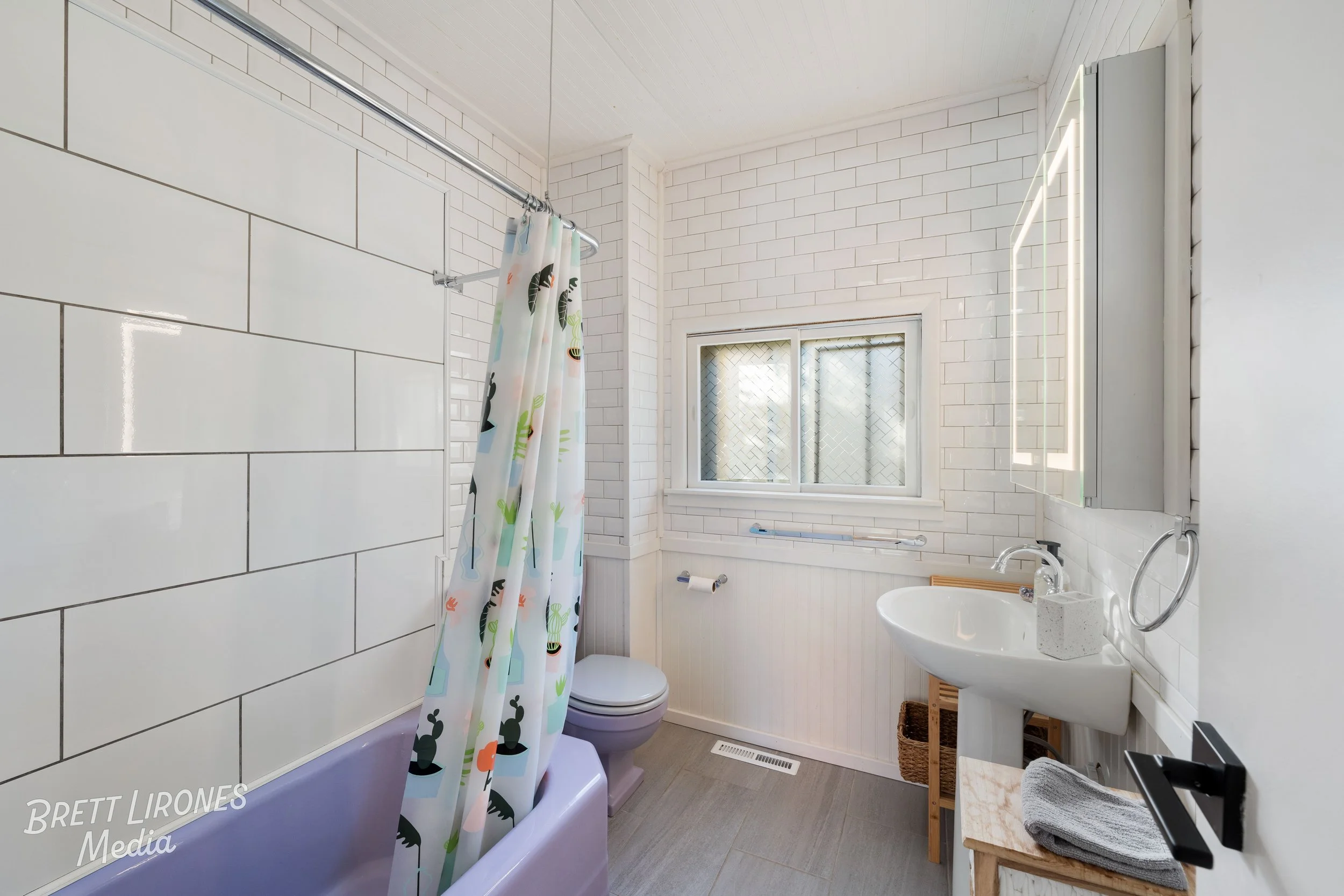 A bright bathroom with white subway tiles, a small window, a purple bathtub with a playful dinosaur shower curtain, a white toilet, a pedestal sink, a mirror with LED lighting, a wooden basket, and a small table with a gray towel.