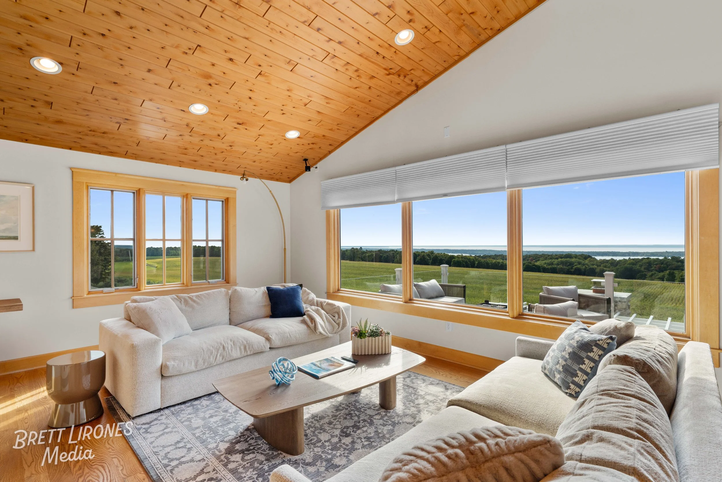 Living room with large windows showing green landscape and water, cream-colored sofas, wooden ceiling, and coffee table.