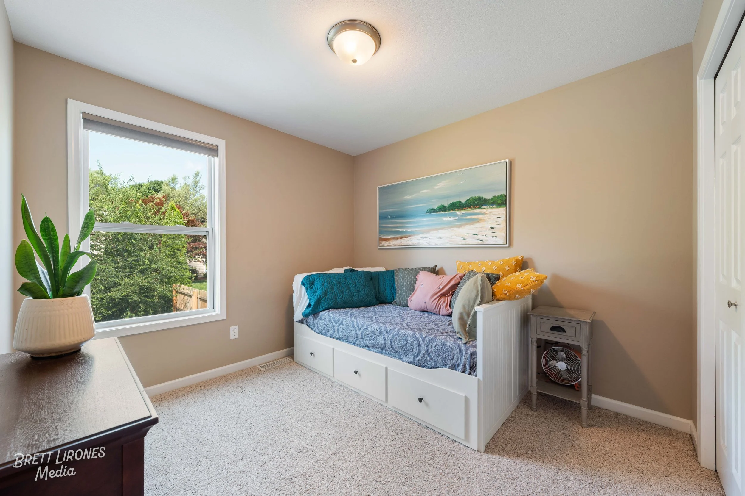A cozy bedroom with a window showing trees and a blue sky, a white daybed with multiple colorful pillows, a landscape painting of a beach scene on the wall, a small gray nightstand with a fan underneath, and a potted plant on a dark wood dresser.