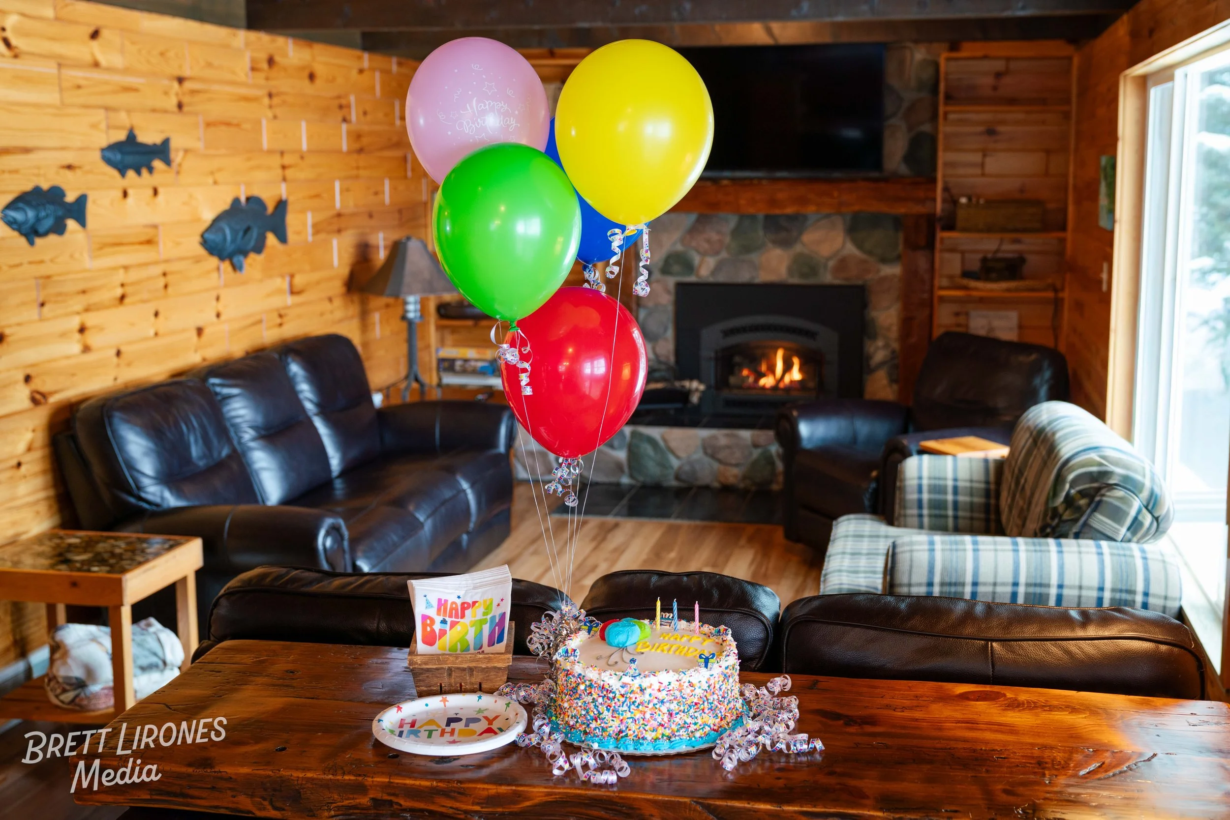 A birthday celebration scene in a cozy living room featuring a cake with colorful sprinkles, birthday candles, and a 'Happy Birthday' decoration on a table. There are five balloons in pink, yellow, green, and red tied together, and party plates with 