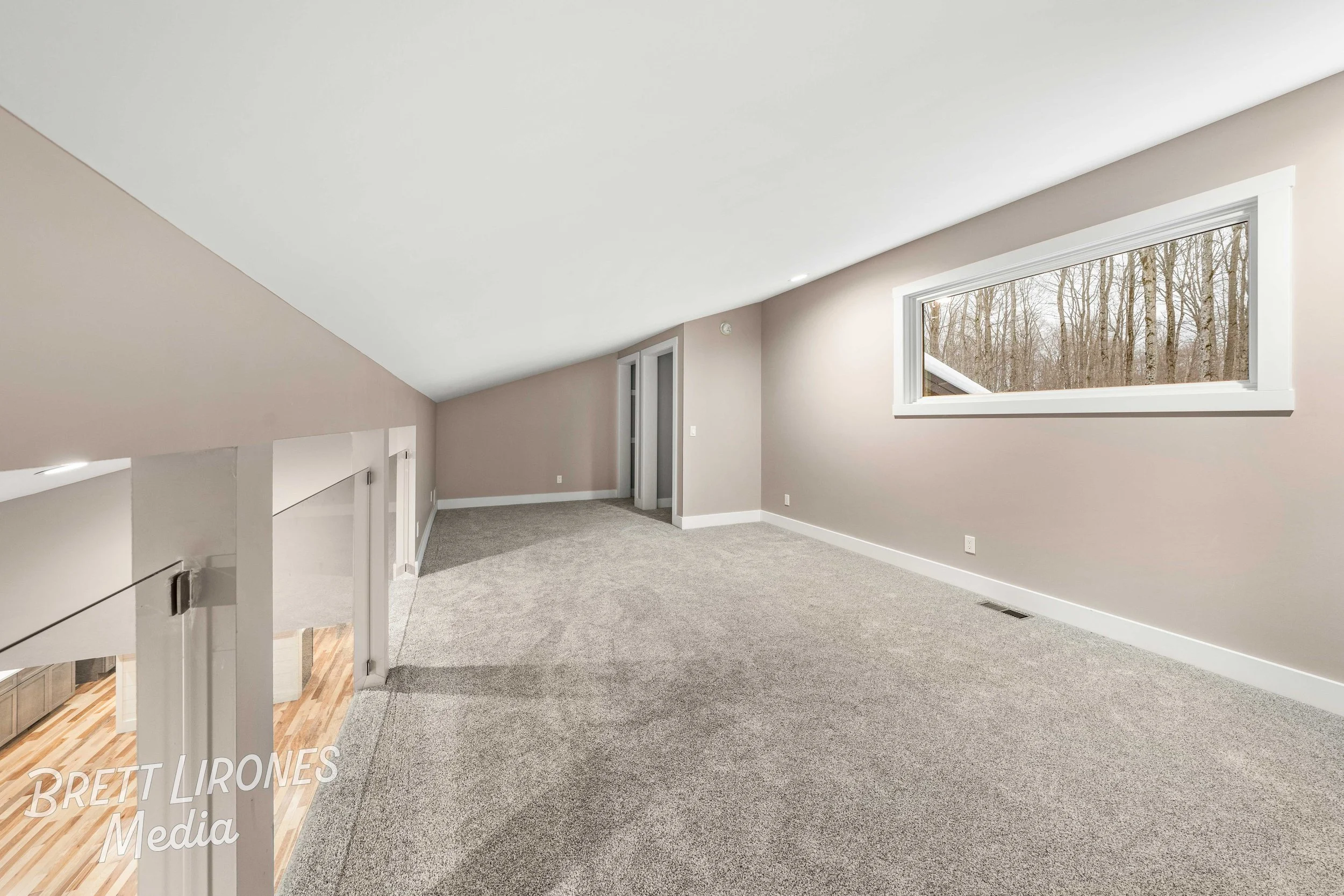 Empty loft with beige walls, gray carpet, a large horizontal window, and a partial view of a staircase with a white railing.