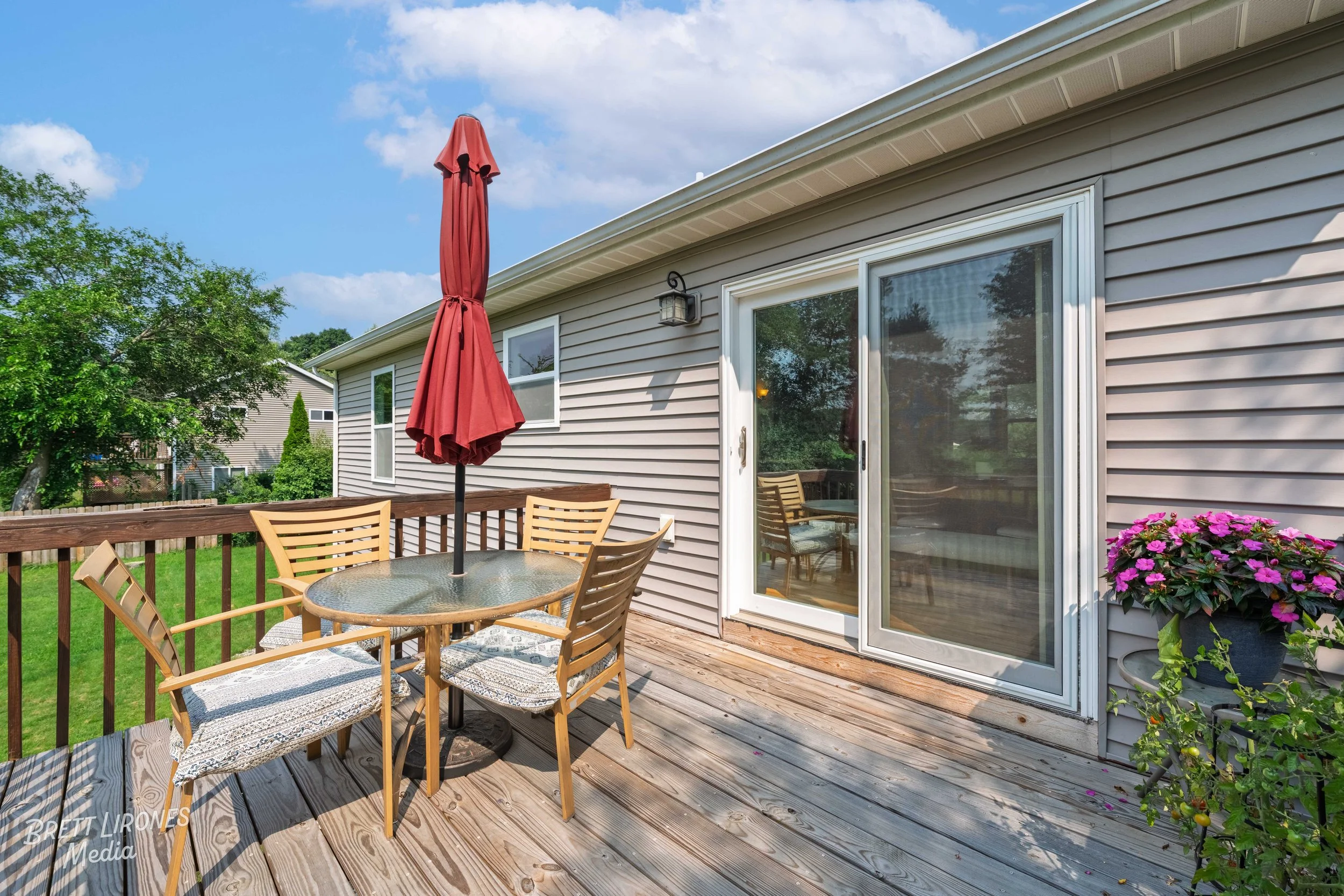 A wooden deck with a glass outdoor table, four wooden chairs with cushions, and a red patio umbrella. The house has beige siding, a sliding glass door, and pink flowers in a pot. The deck is surrounded by green grass and trees, with a neighboring hou