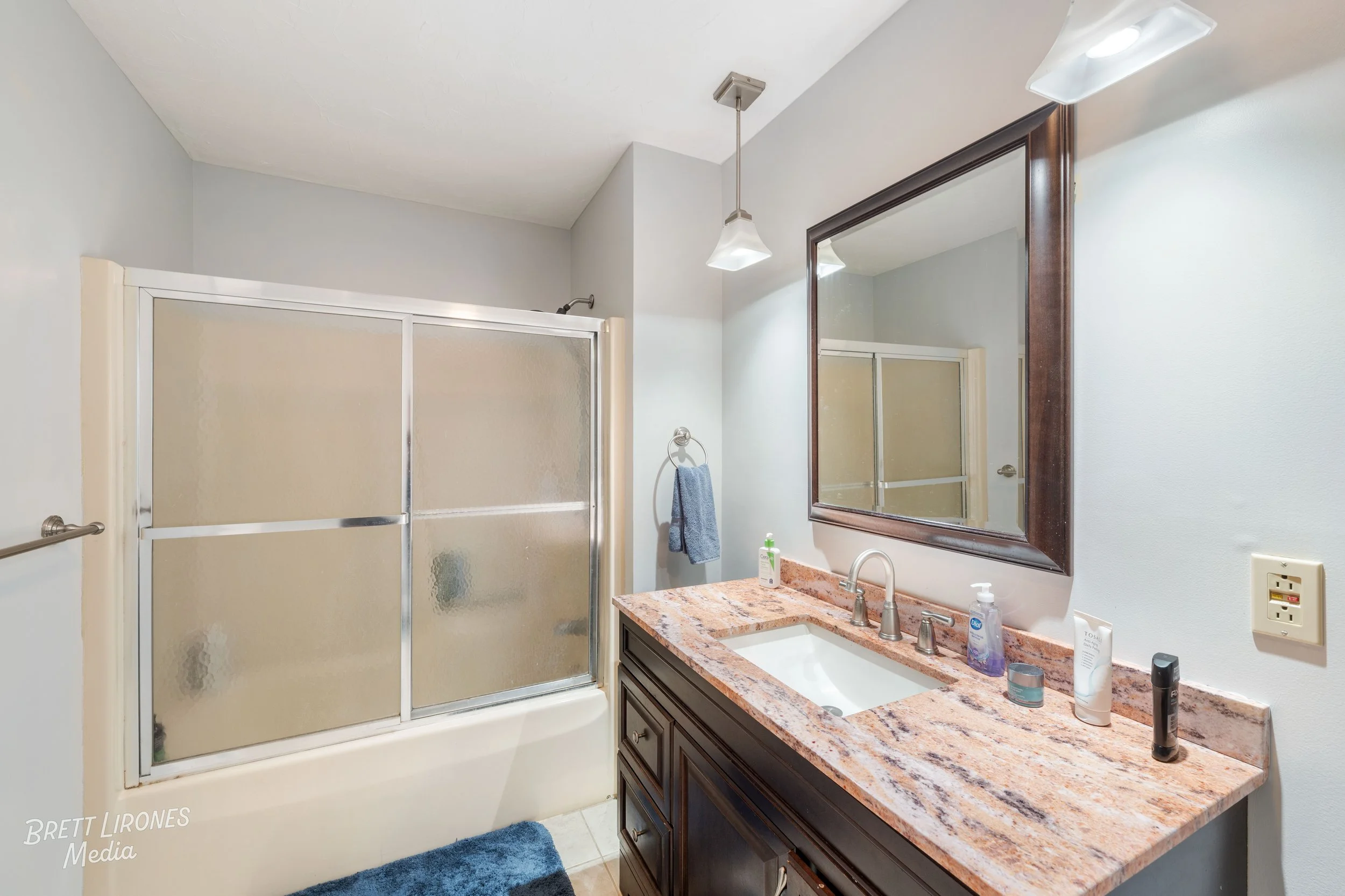 Bathroom with a shower enclosure with frosted glass, a vanity with a granite countertop, a large mirror, and various toiletries on the counter.