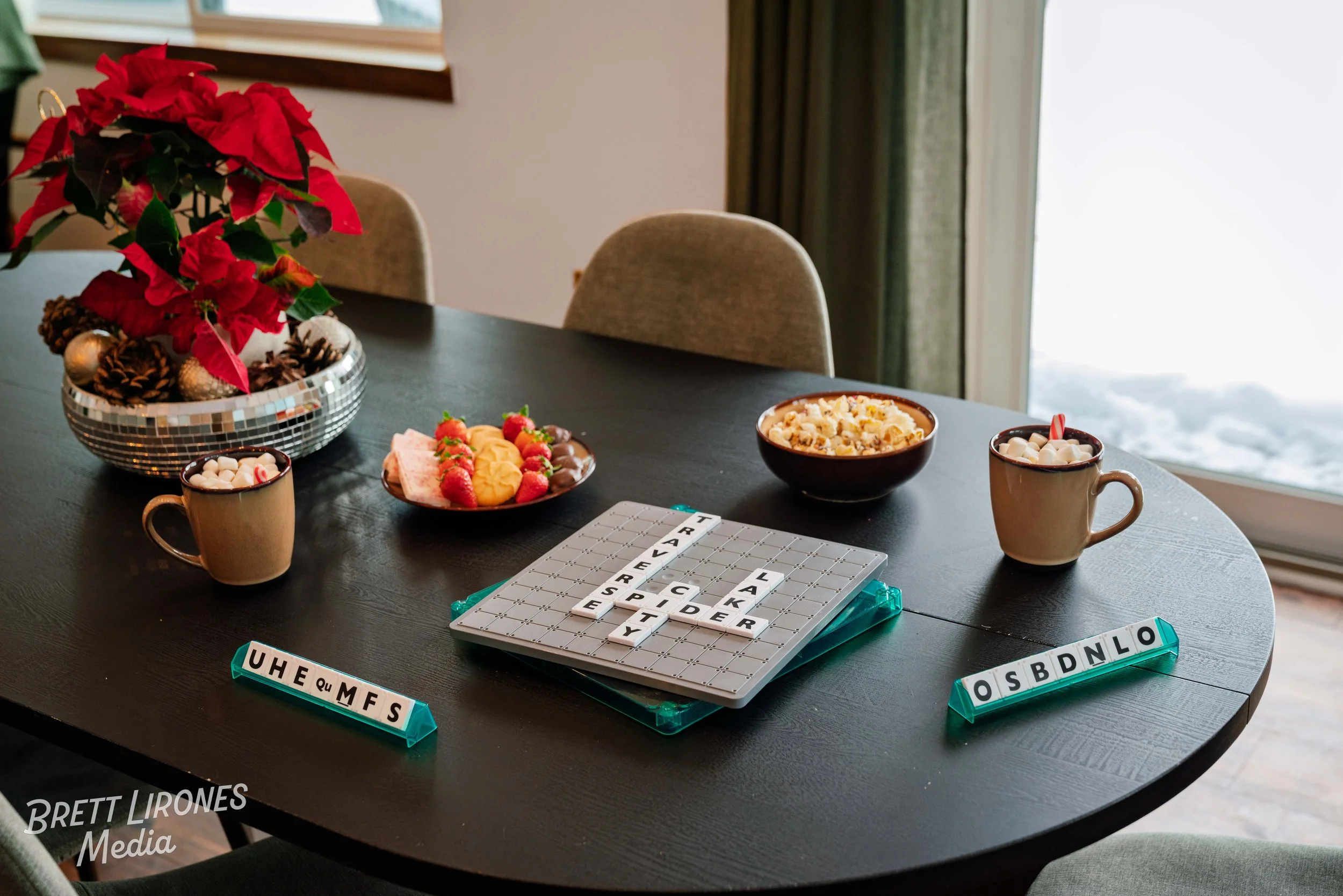 A black oval dining table with holiday decorations, snacks, and board game pieces. There is a poinsettia plant, a bowl of popcorn, a plate of cookies and strawberries, and two mugs with hot chocolate topped with marshmallows. Scrabble game tiles spel