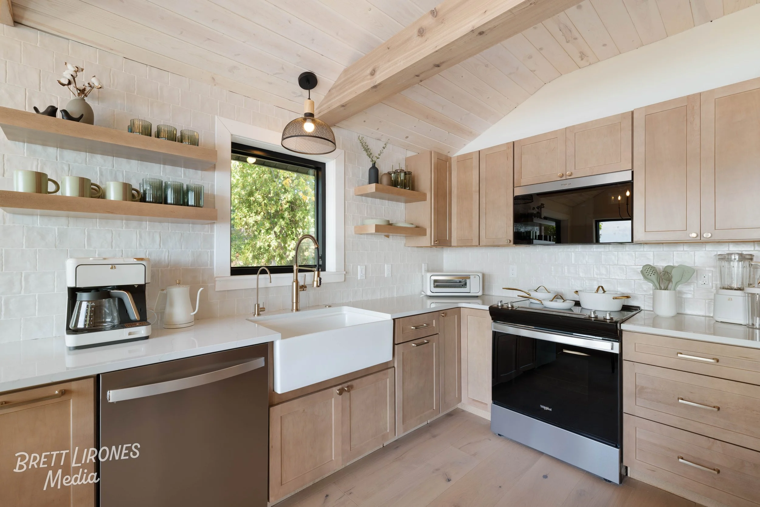 Modern kitchen with light wood cabinets, white subway tile backsplash, farmhouse sink, window above sink, and open shelves with cups and decor. Appliances include a coffee maker and oven. Natural wood ceiling with support beam and pendant light.