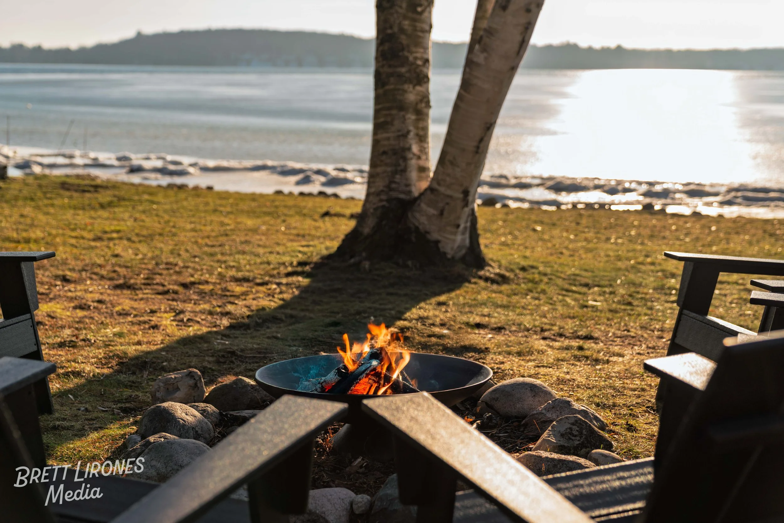 A lakeside scene during sunset with a small campfire surrounded by chairs on the grass, a large tree casting a shadow, and calm water in the background.