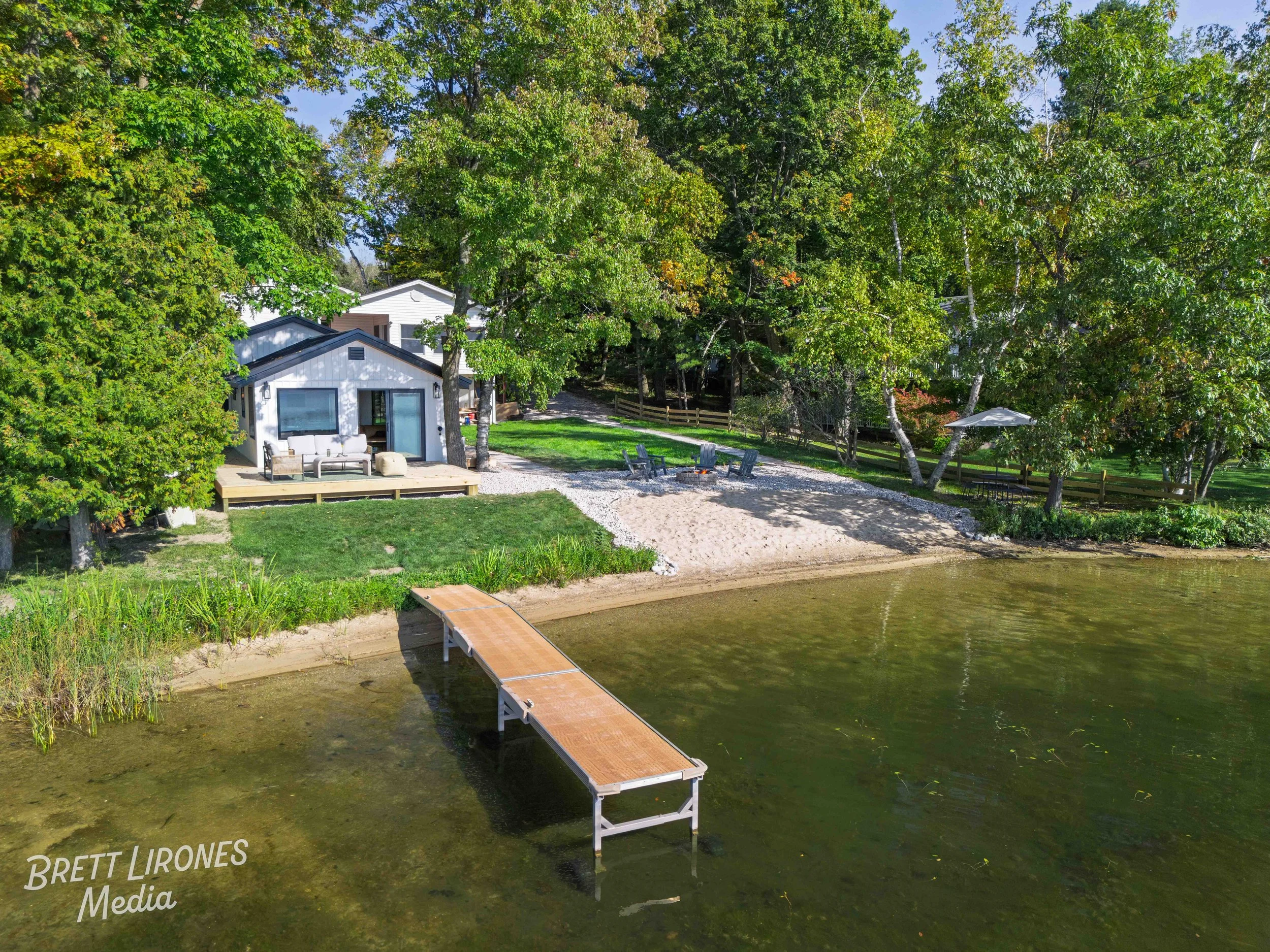 A lakeside backyard with a wooden dock extending into the water, a sandy beach area, and a small house with a deck surrounded by trees.