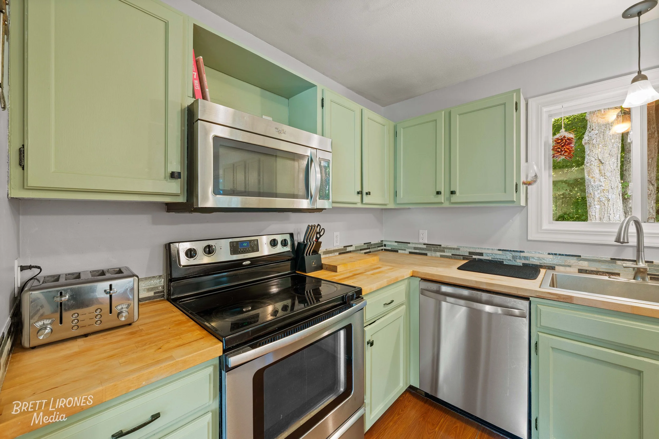 Kitchen with light green cabinets, a stainless steel microwave above a black and stainless steel oven, a stainless steel dishwasher, a wooden countertop, a white window showing outdoors, and a small glass hanging ornament near the window.