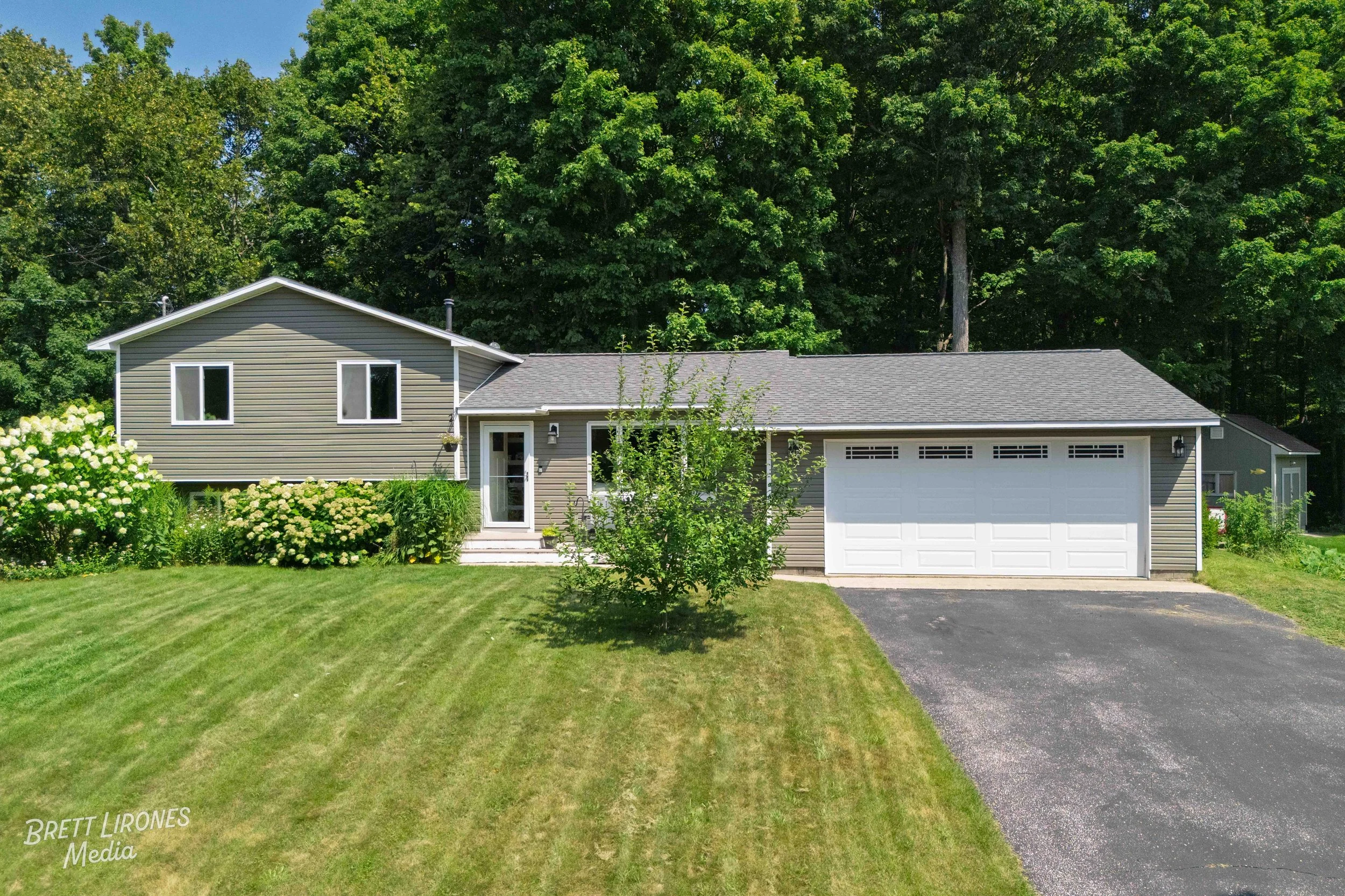 A single-family house with beige siding, white garage door, and a small front porch, surrounded by a well-maintained lawn and lush green trees in the background.