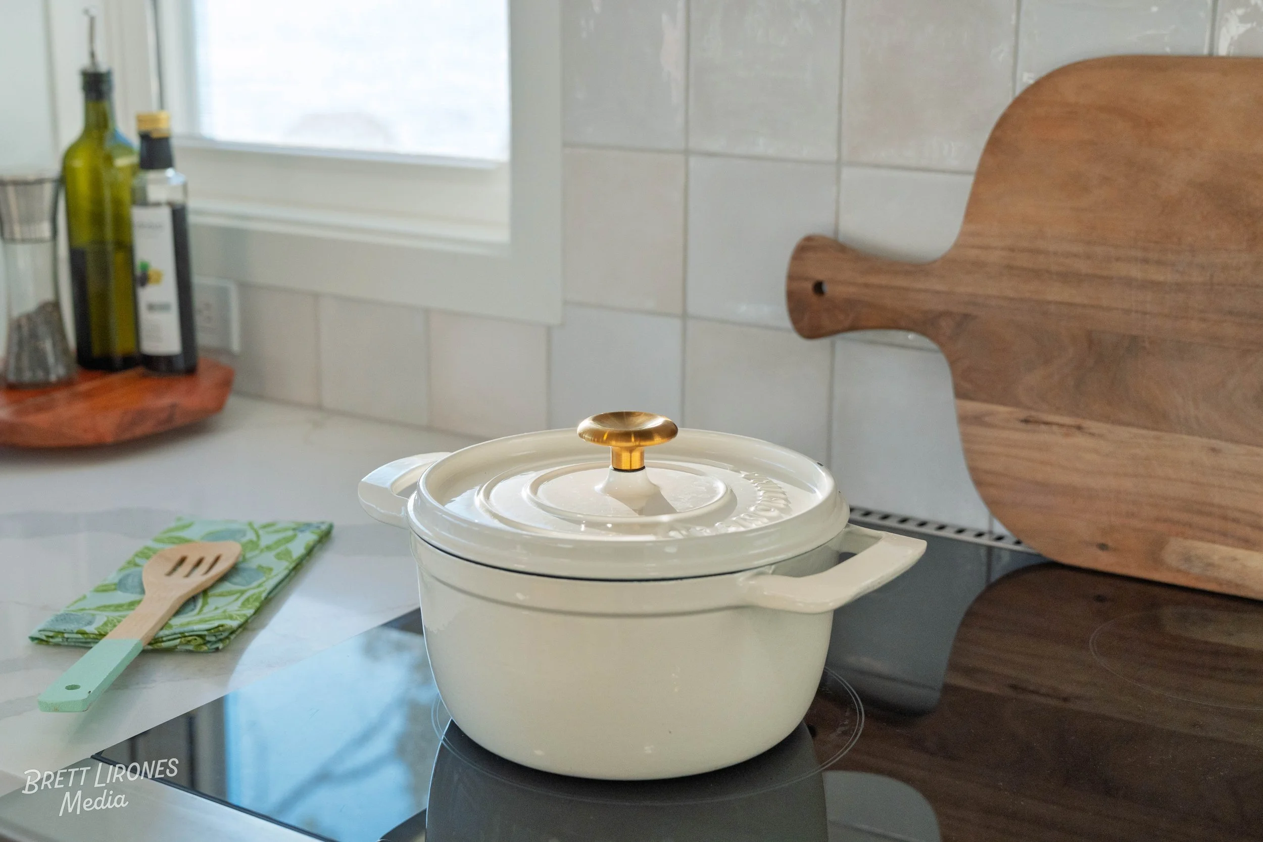 Cream-colored pot with a glass lid and gold handle on a black stovetop in a bright kitchen with white tile backsplash, wooden cutting board, and bottles of oil and vinegar on a wooden shelf.