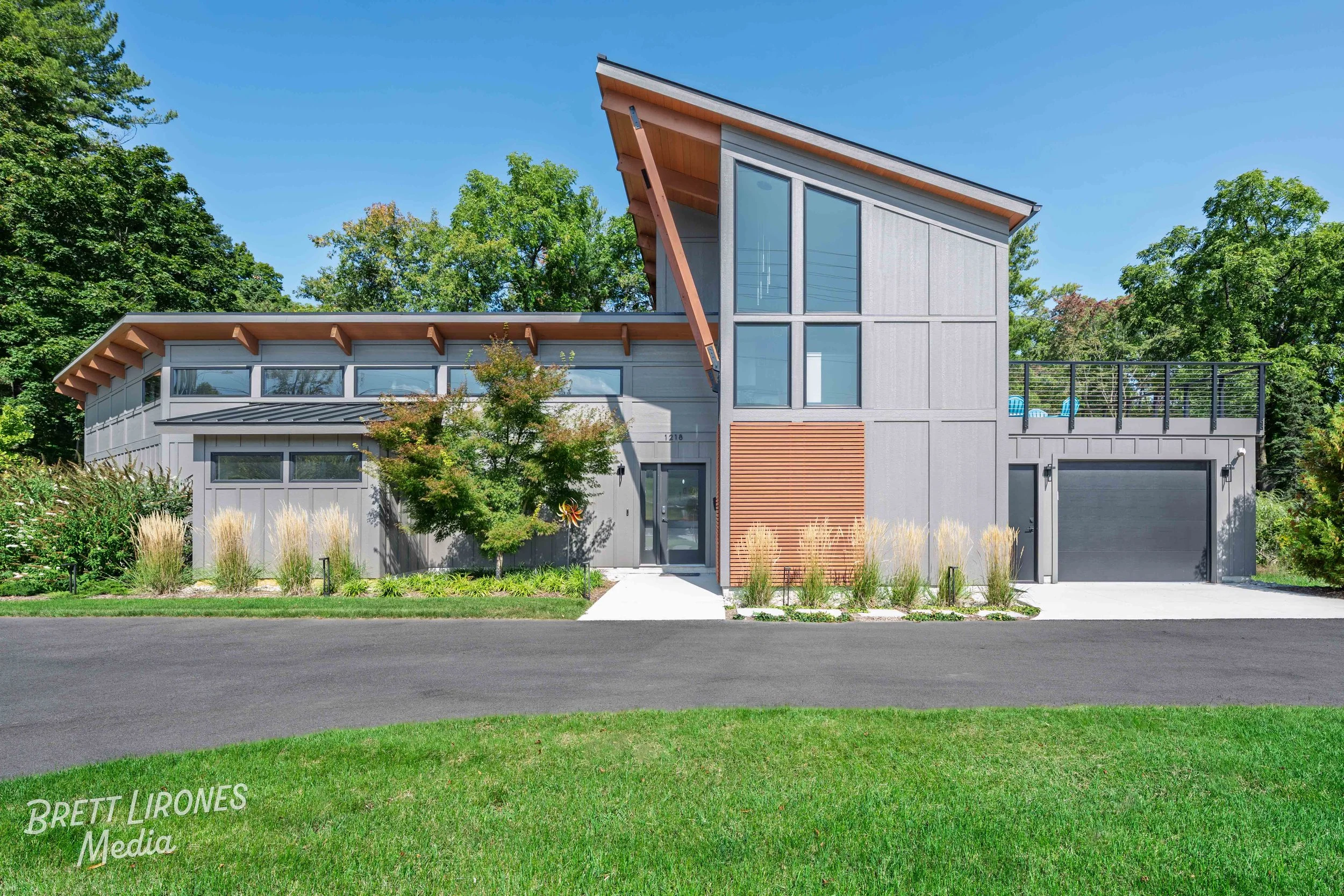 Modern gray house with large windows, a garage, and a landscaped front yard with trees and bushes, under a clear blue sky.