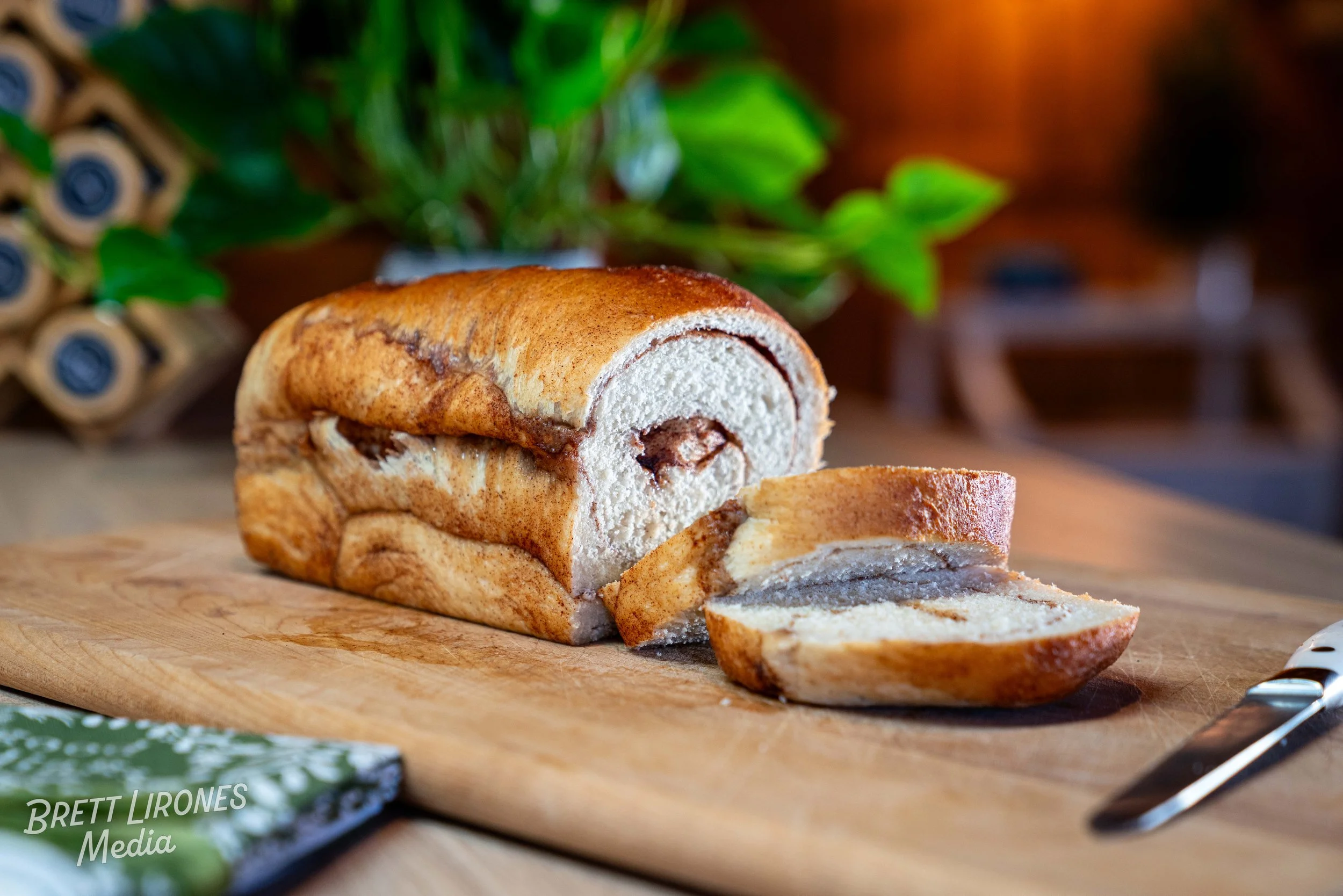 A loaf of cinnamon bread on a wooden cutting board, with a knife and a slice cut from it, in a cozy kitchen setting with green plants in the background.