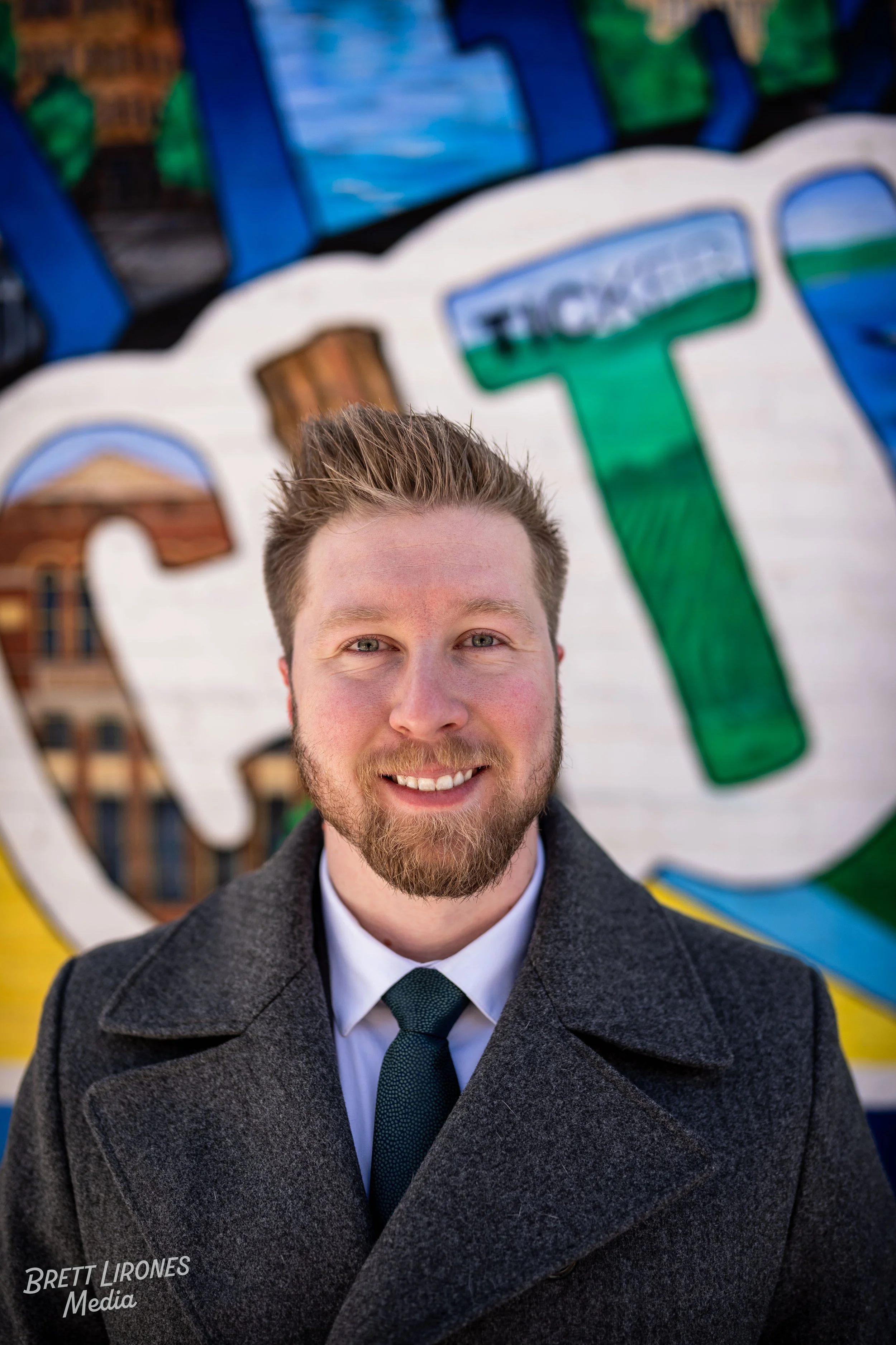 A young man with light skin, a beard, and short, styled hair, dressed in a gray coat and dark tie, smiling at the camera in front of a colorful mural wall.
